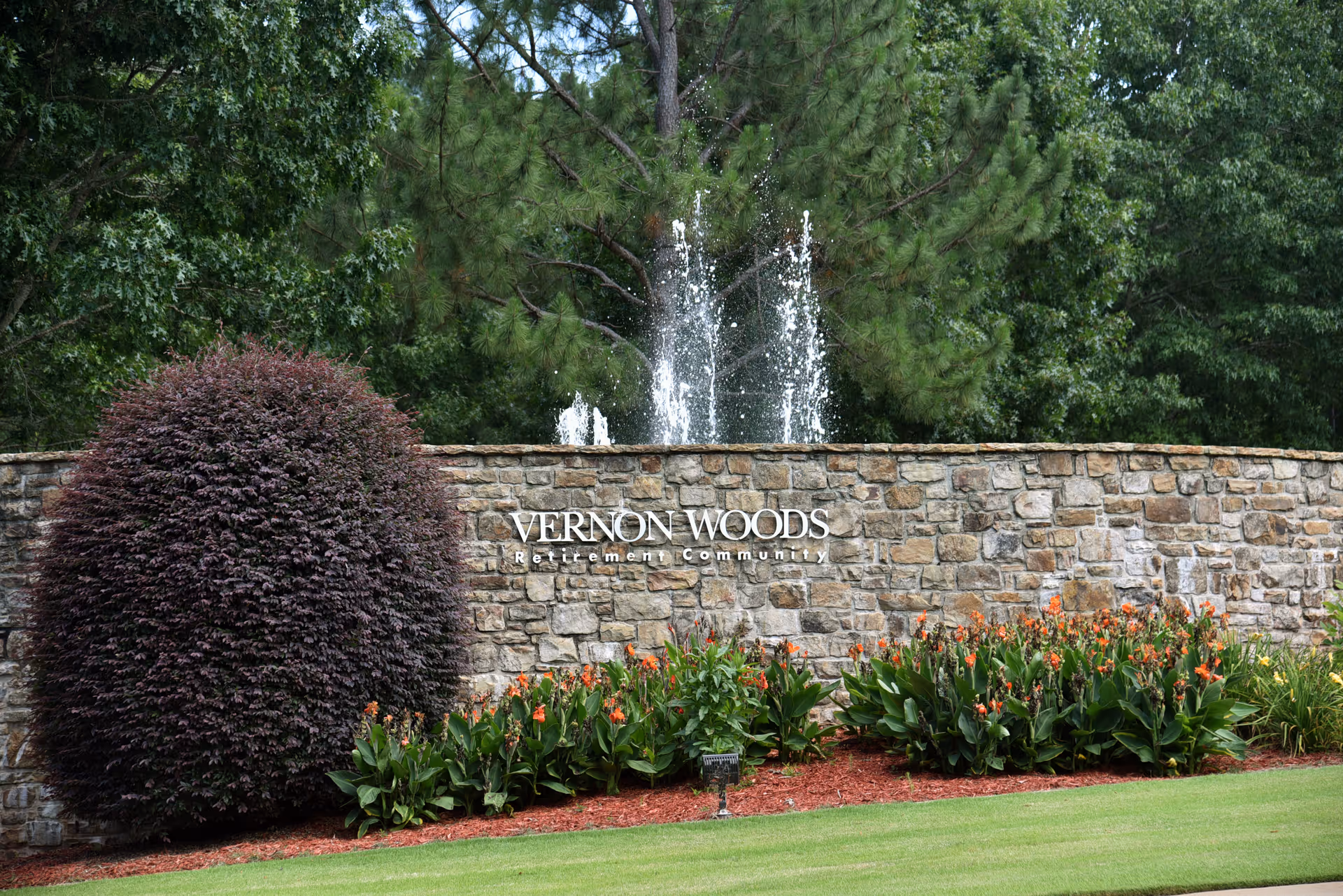 Stone wall with the sign 'VERNON WOODS Retirement Community' in front of a water fountain, surrounded by green trees, bushes, and flowering plants.
