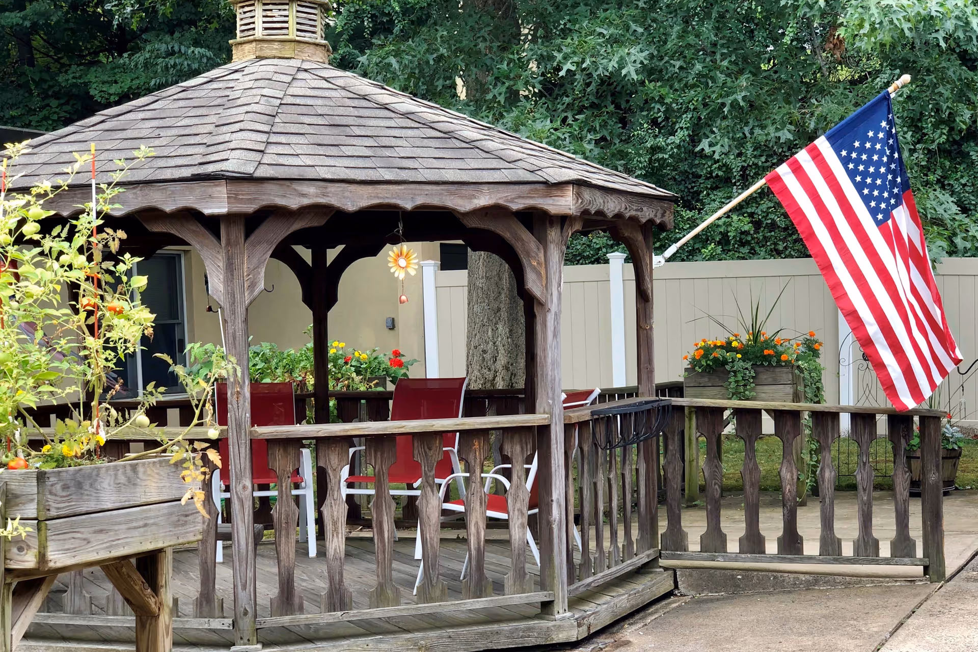 A wooden gazebo with a shingled roof in an outdoor garden area. Inside the gazebo are red chairs and a hanging decorative flower. There are planters with flowers and greenery around the gazebo, and an American flag is mounted on the right side. A beige fence and trees are visible in the background.