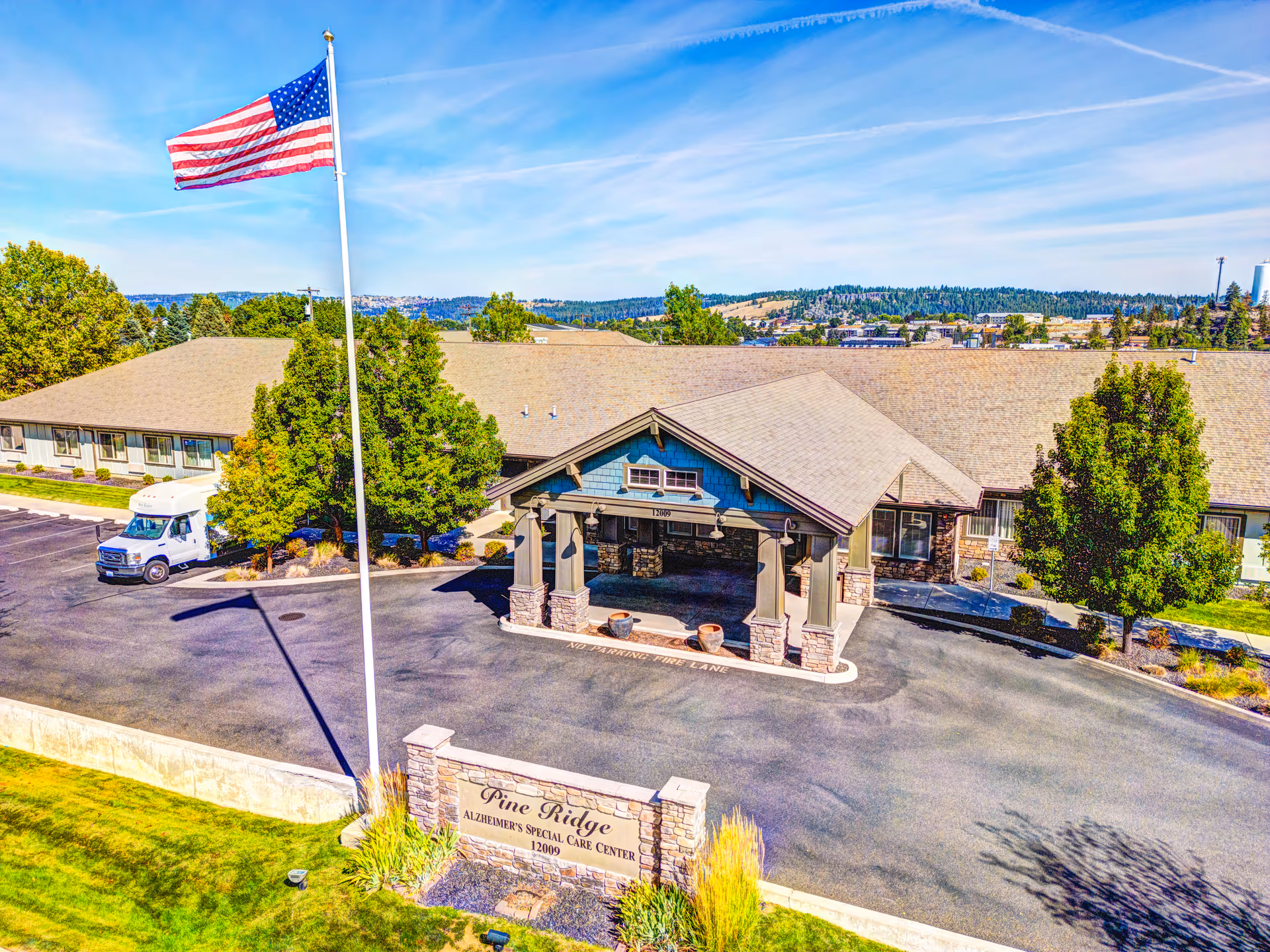 Exterior view of Pine Ridge Alzheimer's Special Care Center, showing a single-story building with a covered entrance, an American flag on a tall flagpole, a parking area with a white van, and surrounding trees and greenery under a blue sky.