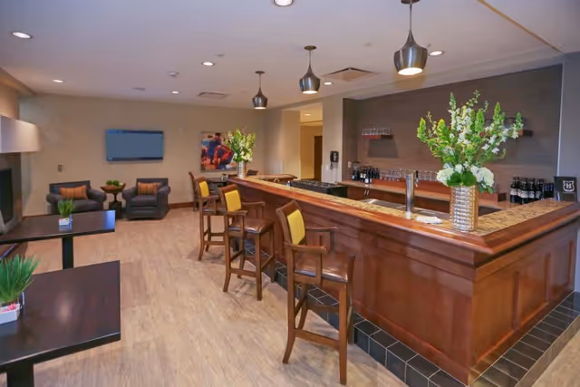 Well-lit lounge area with a wooden bar counter, barstools, seating, and floral arrangements.