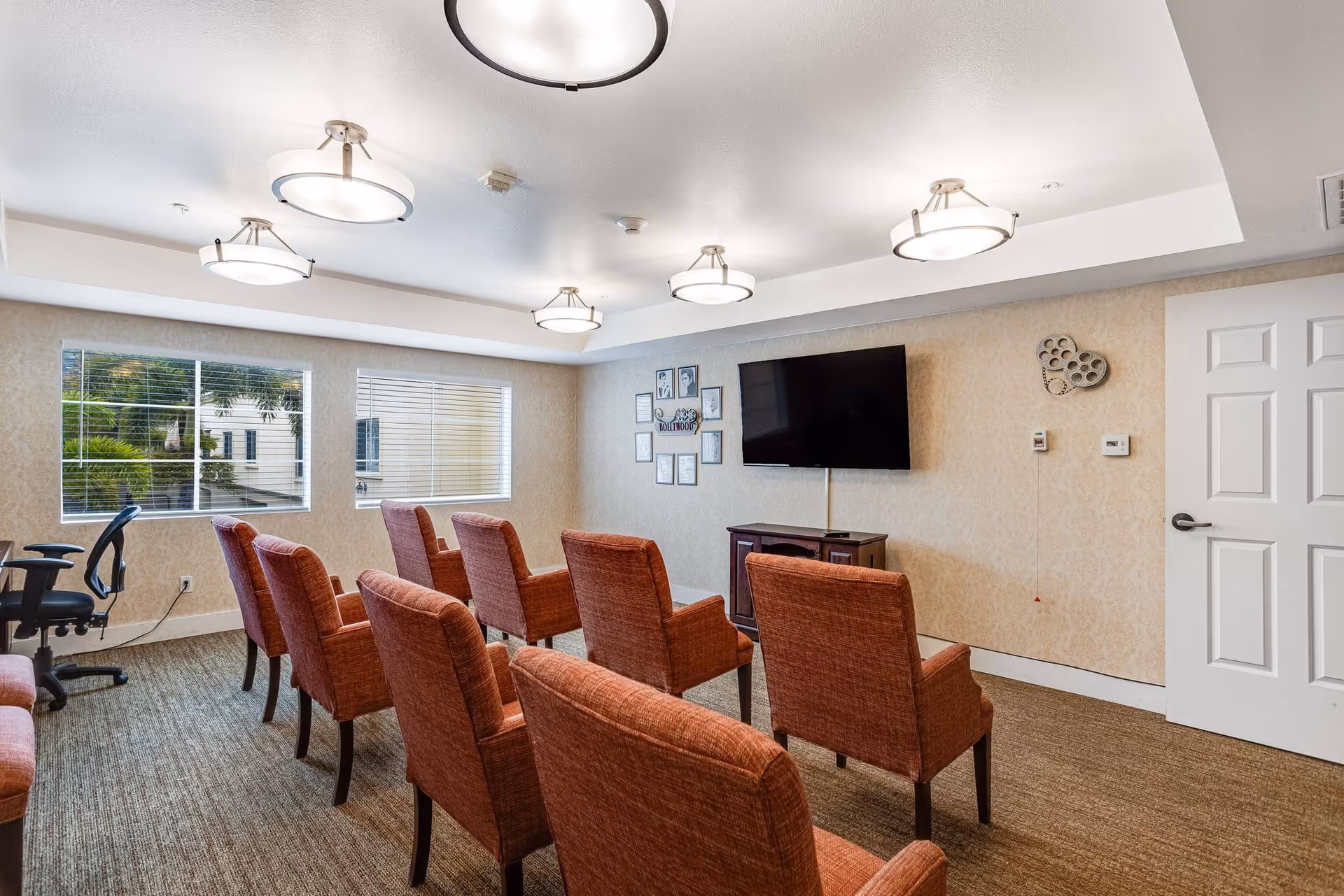 Small media room with rows of red upholstered chairs facing a wall-mounted TV and windows along the left wall.