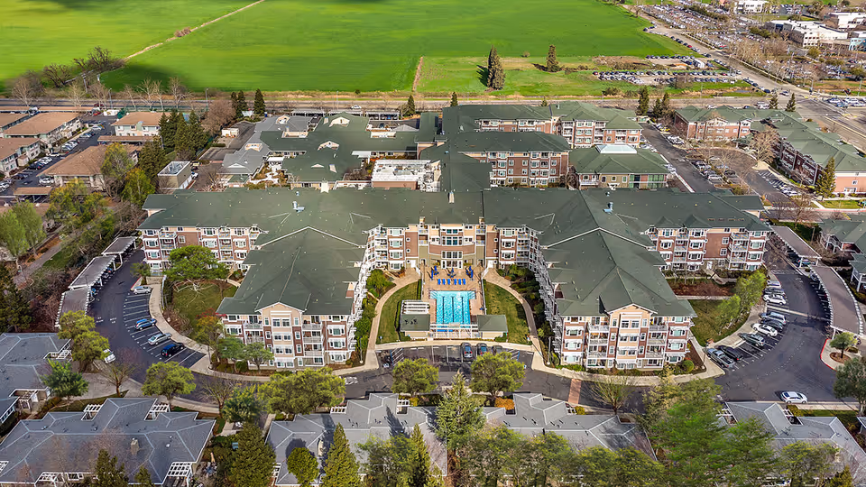 Aerial view of University Retirement Community showing a large multi-wing building with green roofs surrounding a central courtyard that features a swimming pool and seating areas. The complex is surrounded by parking lots, trees, and open green fields in the background.