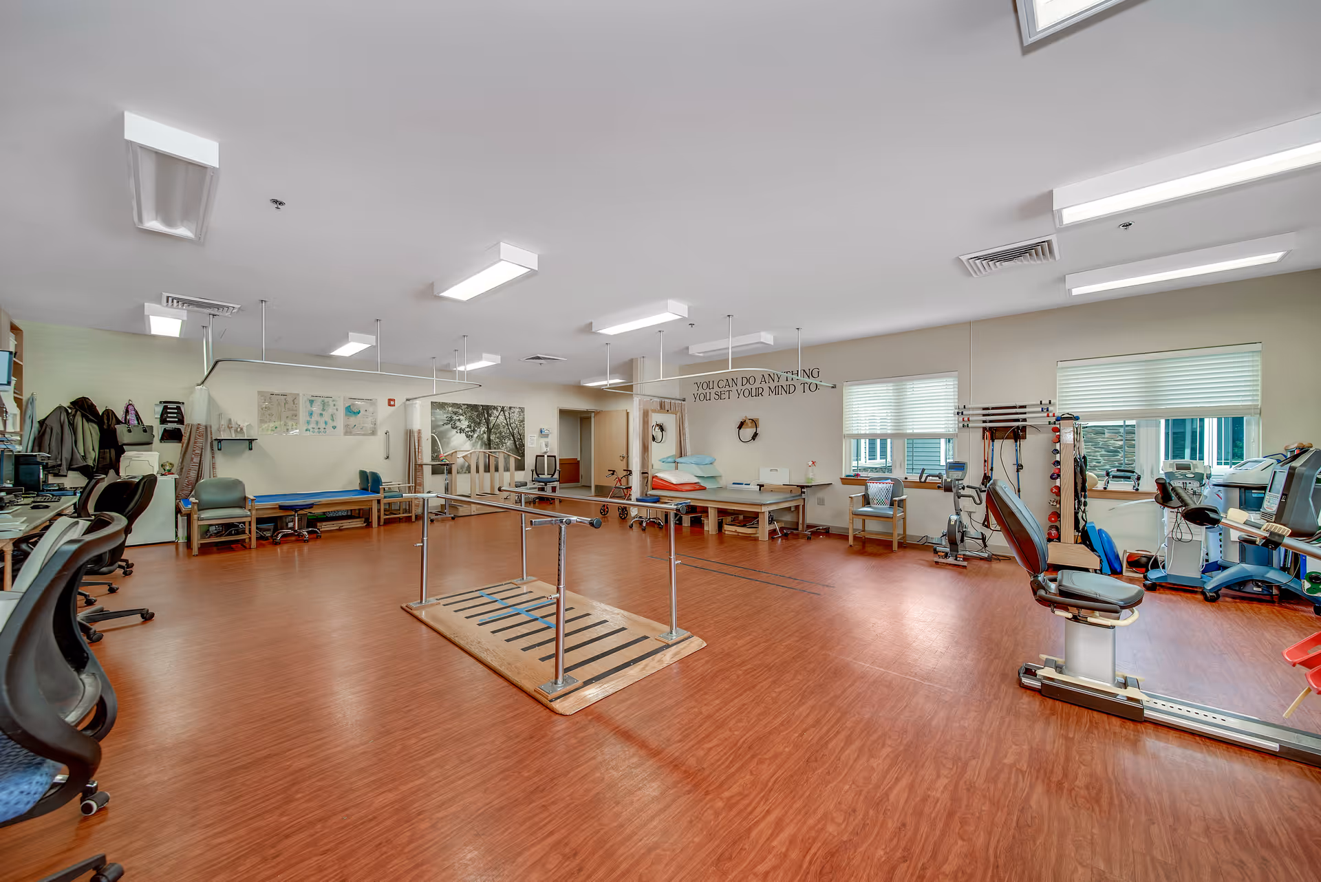 A spacious physical therapy or rehabilitation room with wooden flooring, parallel bars for walking exercises in the center, exercise equipment including stationary bikes and treadmills along the right side, chairs and benches along the walls, and motivational text on the wall that reads 'YOU CAN DO ANYTHING YOU SET YOUR MIND TO'. The room is well-lit with ceiling lights and has windows with blinds.