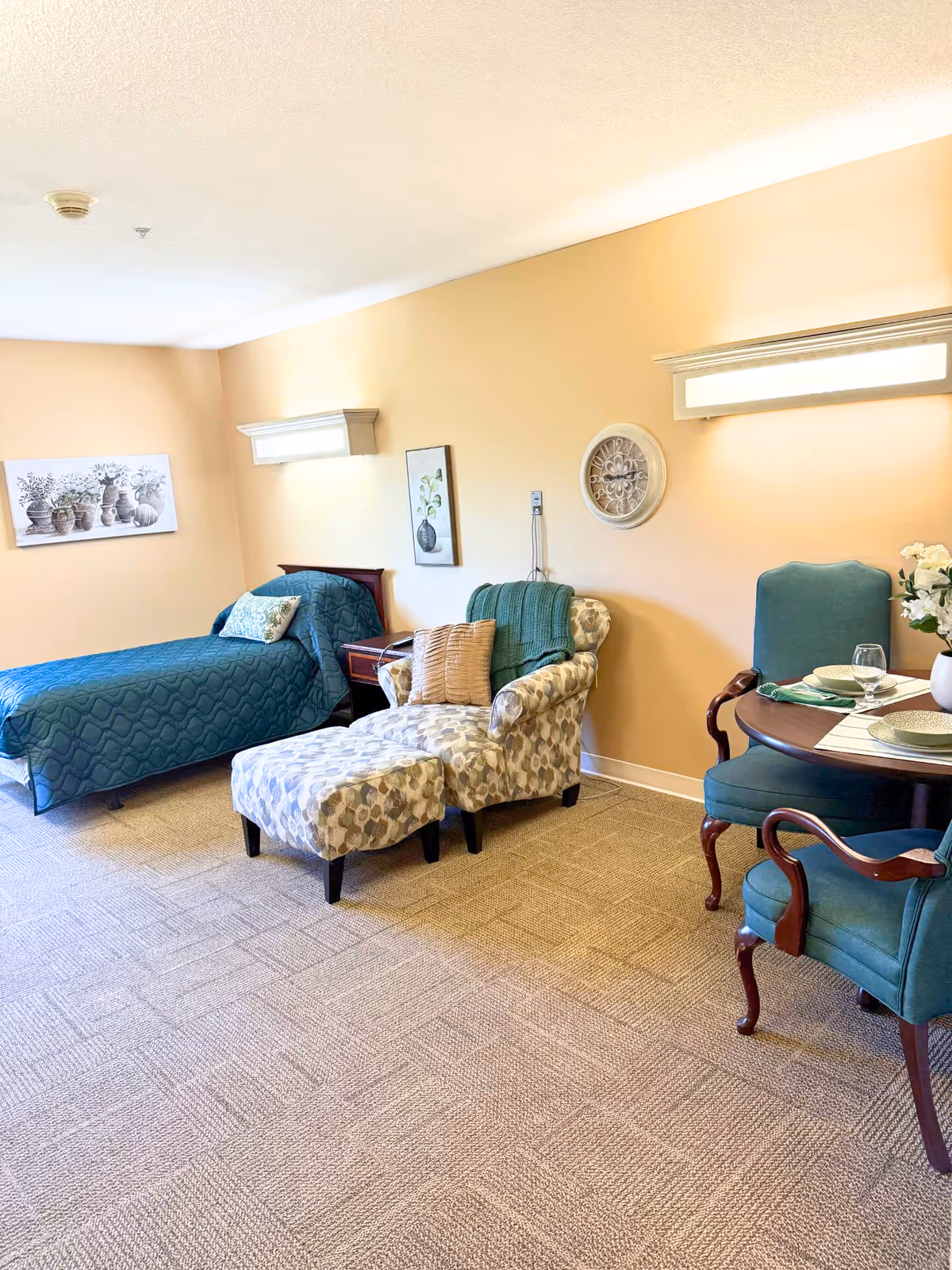 A cozy assisted living room with a single bed covered in a blue quilt, a patterned armchair with a matching ottoman, and a small dining table set with two green upholstered chairs and place settings. The walls are painted beige and decorated with framed artwork and a decorative wall piece. The room is well-lit with wall-mounted lights.