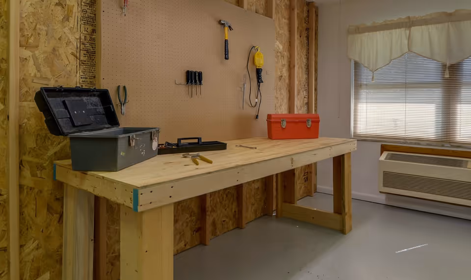 A small workshop interior with a wooden workbench, toolboxes, hand tools on a pegboard, and a window with an air conditioning unit.