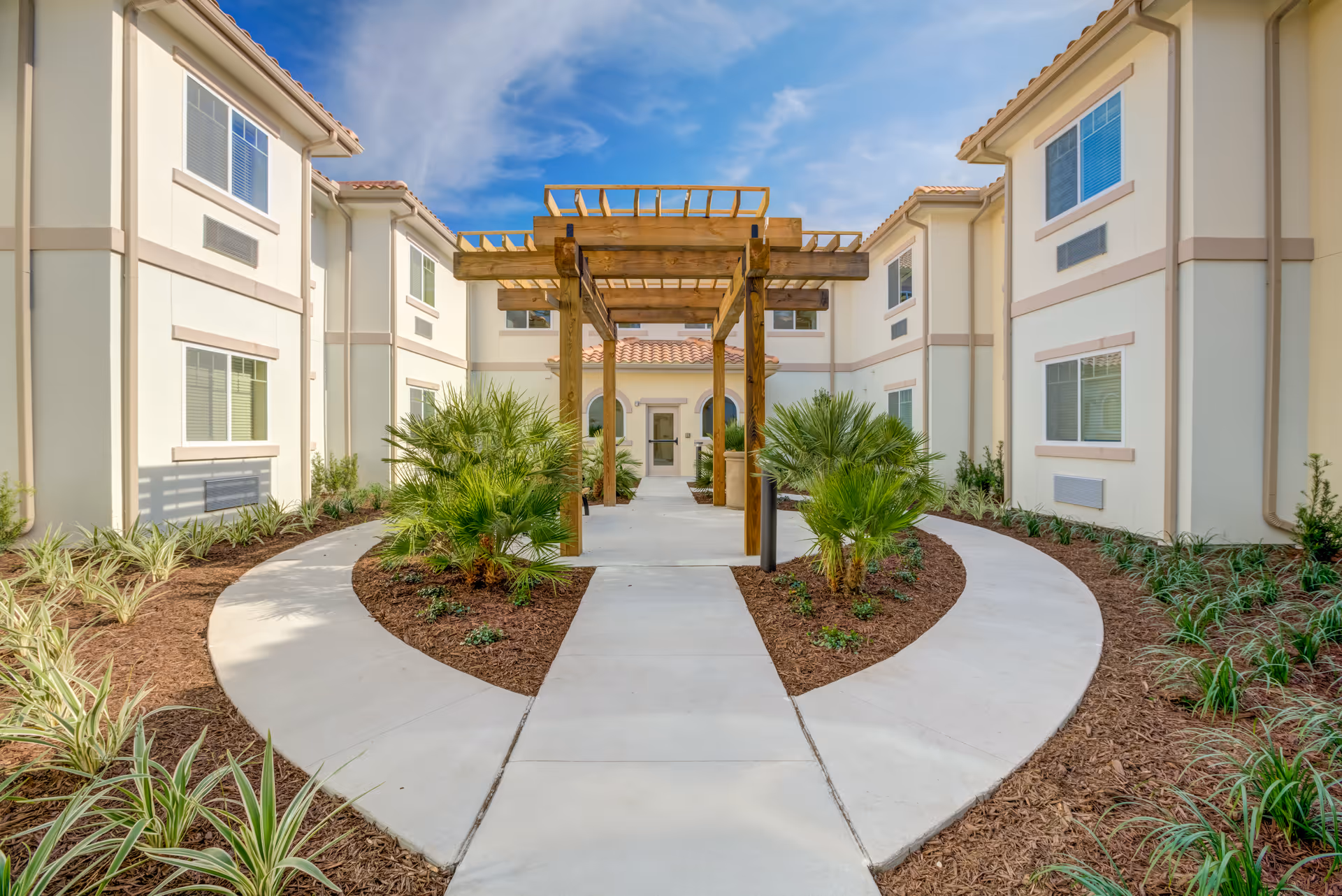 Curved concrete walkway leads through landscaped beds to a wooden pergola and main entrance between two-story cream-colored buildings.