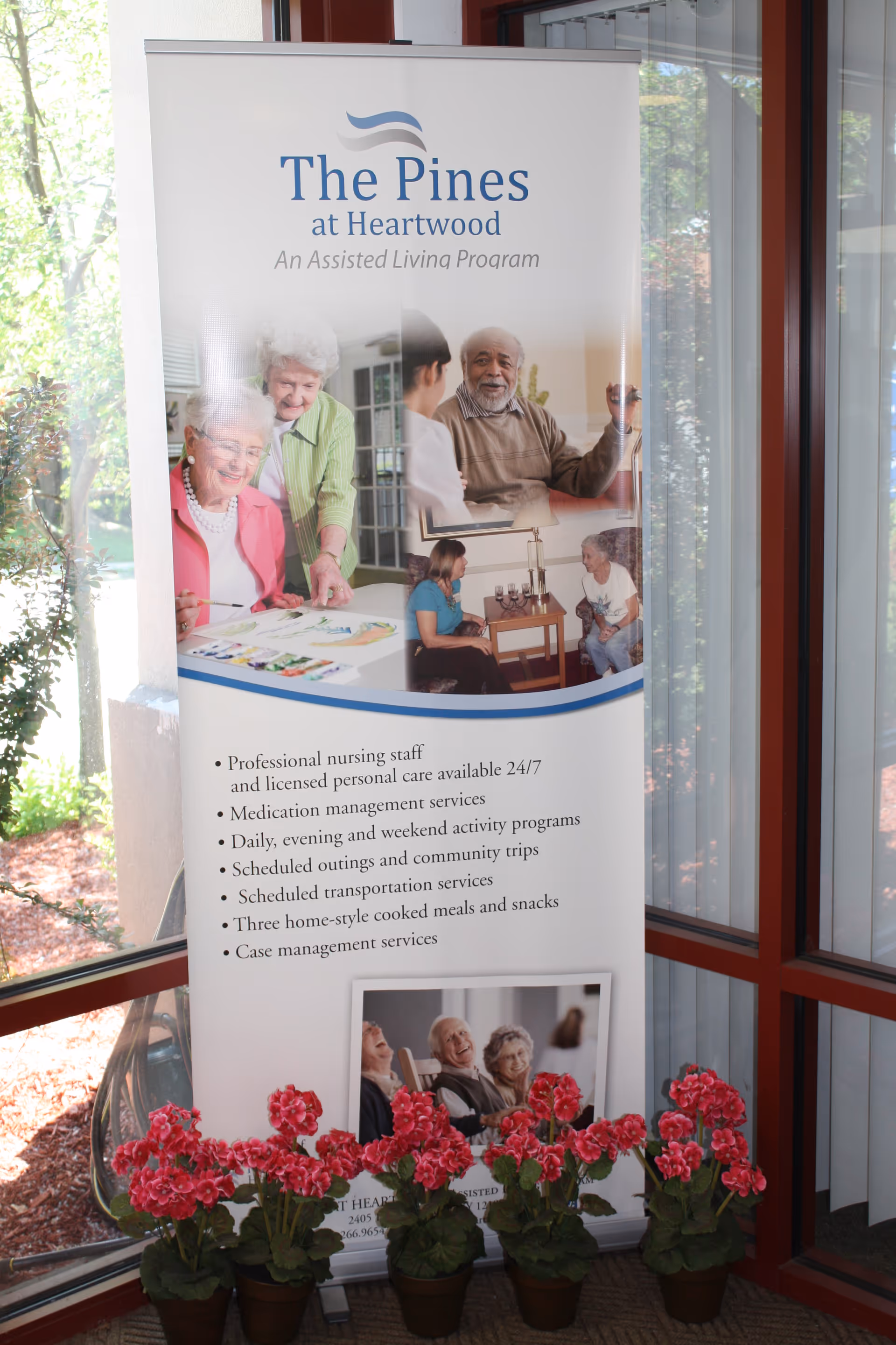 A vertical banner for The Pines at Heartwood, an assisted living program, displayed indoors near a window. The banner features images of elderly people engaging in activities and socializing. It lists services such as professional nursing staff, medication management, activity programs, transportation, home-style meals, and case management. In front of the banner are five pots of red flowering plants.