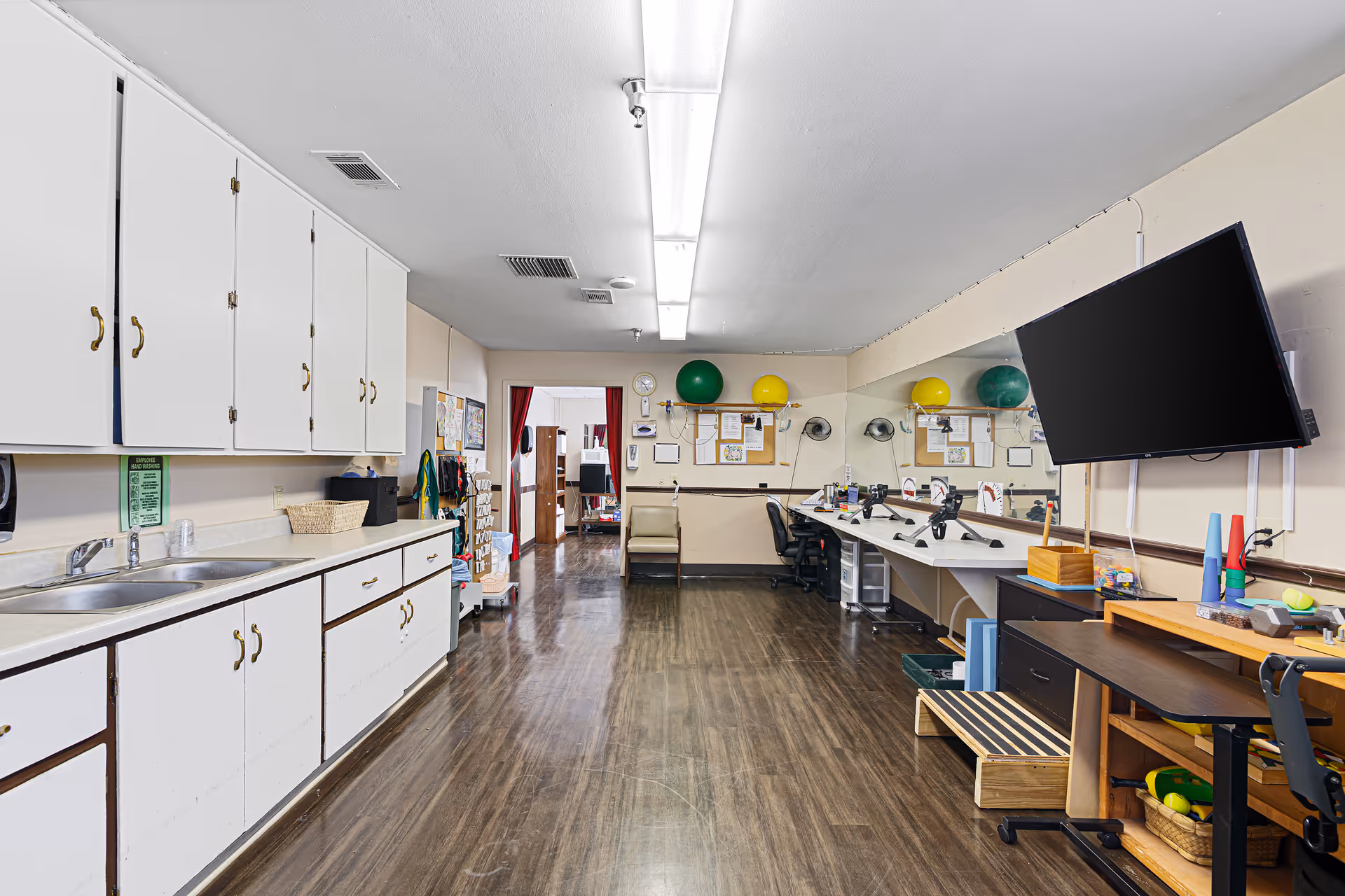 Interior therapy/activity room with cabinets and sink on the left, workstations, equipment and a wall-mounted TV on the right.