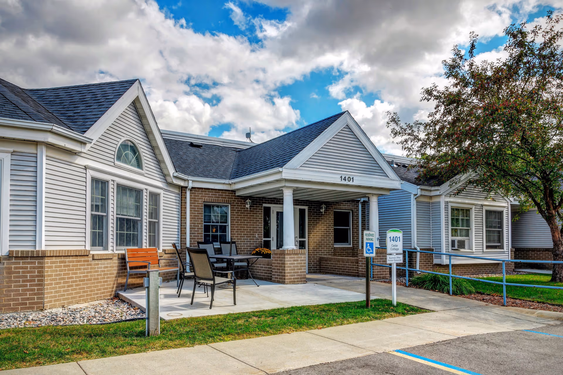 Exterior view of a single-story senior living facility building with a covered entrance, outdoor seating area with chairs and a table, a bench, and a tree on the right side. The building has a combination of brick and siding with multiple windows and a ramp for accessibility. The sky is partly cloudy.