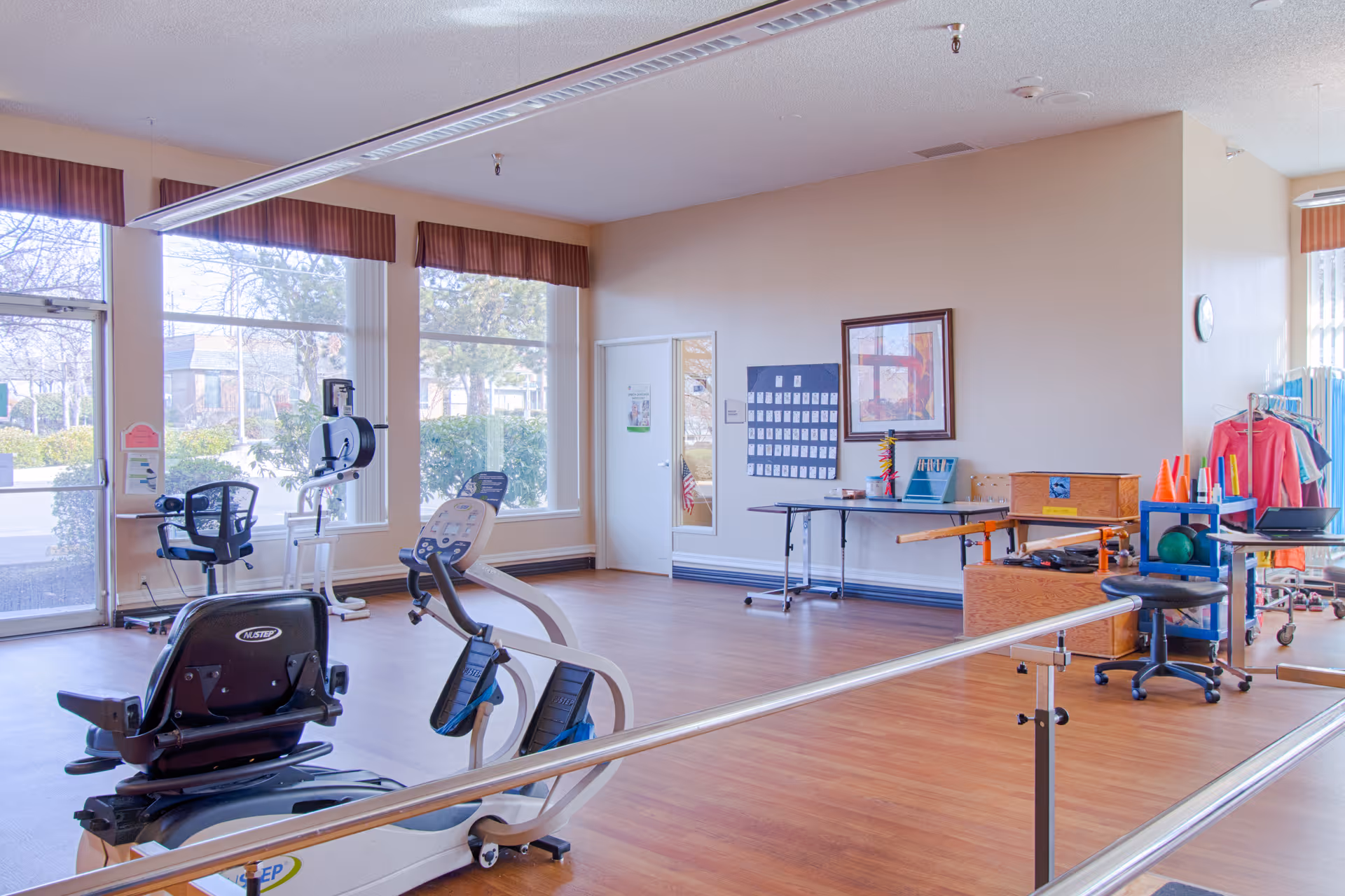 A bright rehabilitation or physical therapy room with large windows letting in natural light. The room contains exercise equipment including a stationary recumbent bike, parallel bars for walking support, and various therapy tools and supplies on tables and shelves. The floor is wooden, and there are some chairs and a rack with clothes in the background.
