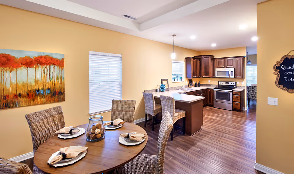 A bright and cozy dining area and kitchen in a senior living facility. The dining area features a round wooden table set with four place settings, each with a plate, napkin, and napkin ring. Four wicker chairs surround the table. The kitchen has dark wooden cabinets, a stainless steel stove, microwave, and a kitchen island with two wicker bar stools. The walls are painted a warm yellow, and there is a colorful painting of trees on the wall near the dining table. A small chalkboard sign is visible on the right wall.