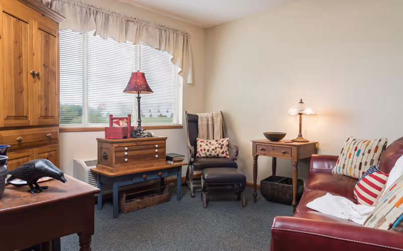 A cozy living room with a red leather sofa adorned with patterned pillows and a white throw blanket. There is a wooden side table with a lamp and a bowl, a dark wooden rocking chair with a matching footrest and a pillow, and a wooden cabinet near a window with blinds and a valance. The room has a carpeted floor and neutral-colored walls.