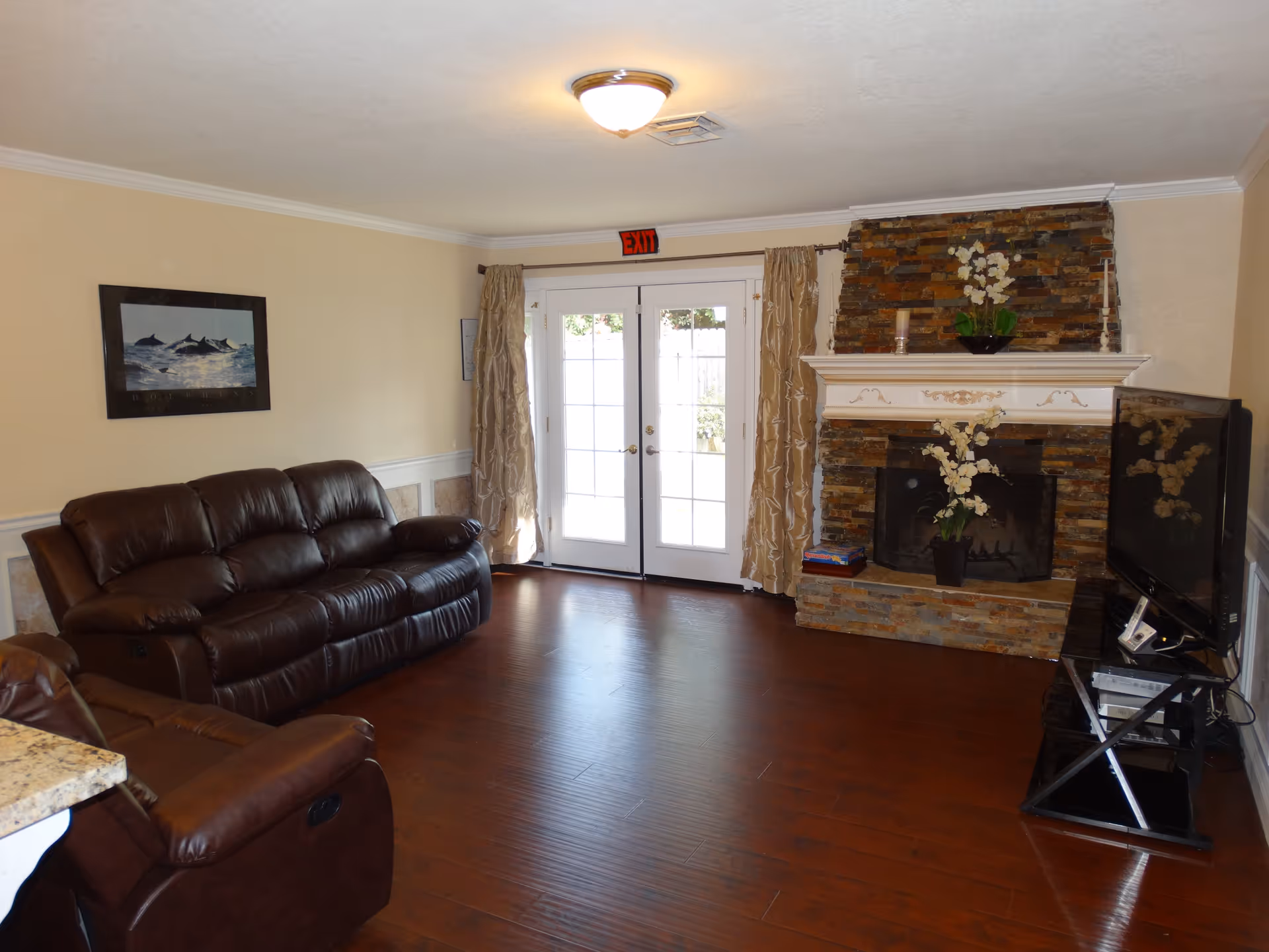 A cozy living room with two brown leather sofas, a stone fireplace decorated with white flowers, a flat-screen TV on a stand, and double glass doors with beige curtains leading outside. The room has wooden flooring and light-colored walls with a dolphin picture hanging on one wall.