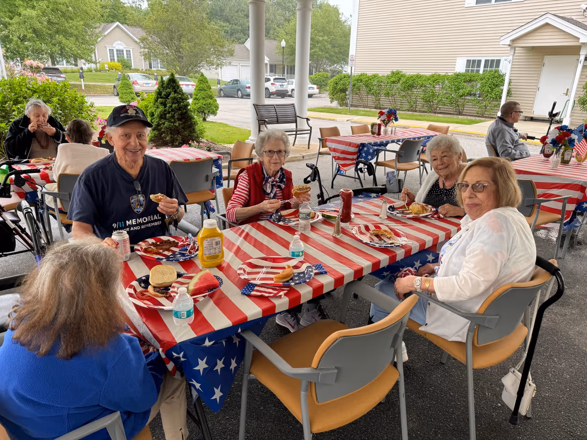 A group of elderly people sitting around a table covered with a red, white, and blue American flag-themed tablecloth, enjoying a meal outdoors under a covered area. The table has plates with food including hot dogs and watermelon, condiments like mustard and ketchup, and bottled water. In the background, there are more tables with similar decorations and a few more people seated. The setting appears to be a communal outdoor dining area at a senior living facility.