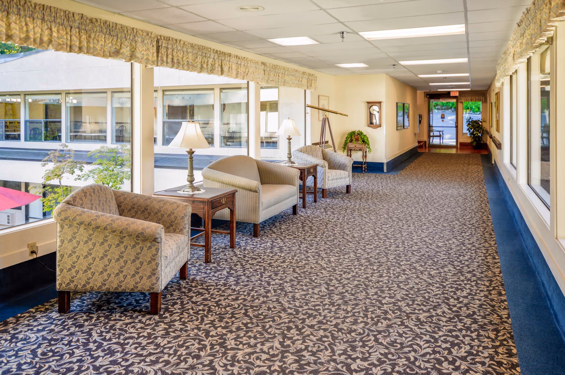 Bright carpeted interior hallway in an assisted living facility with chairs, side tables, lamps and large windows.