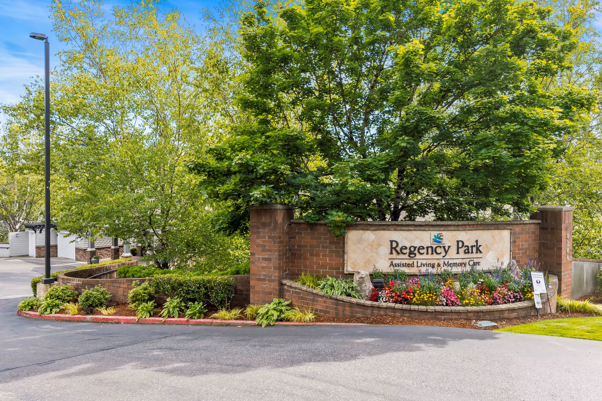 Entrance sign for Regency Park Assisted Living & Memory Care surrounded by green trees, shrubs, and colorful flowers with a paved driveway in front.