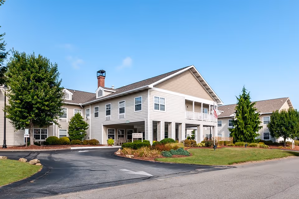 Exterior view of a two-story senior living facility building with beige siding, multiple windows, a covered entrance, and landscaped grounds with trees and shrubs under a clear blue sky.