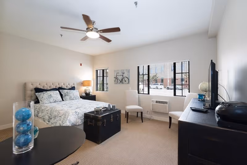 A bright and clean bedroom in a senior living facility featuring a bed with patterned bedding, two chairs near windows with blinds, a ceiling fan, a black trunk at the foot of the bed, a dresser with a TV and decorative items, and a small table with a glass vase containing blue decorative balls.