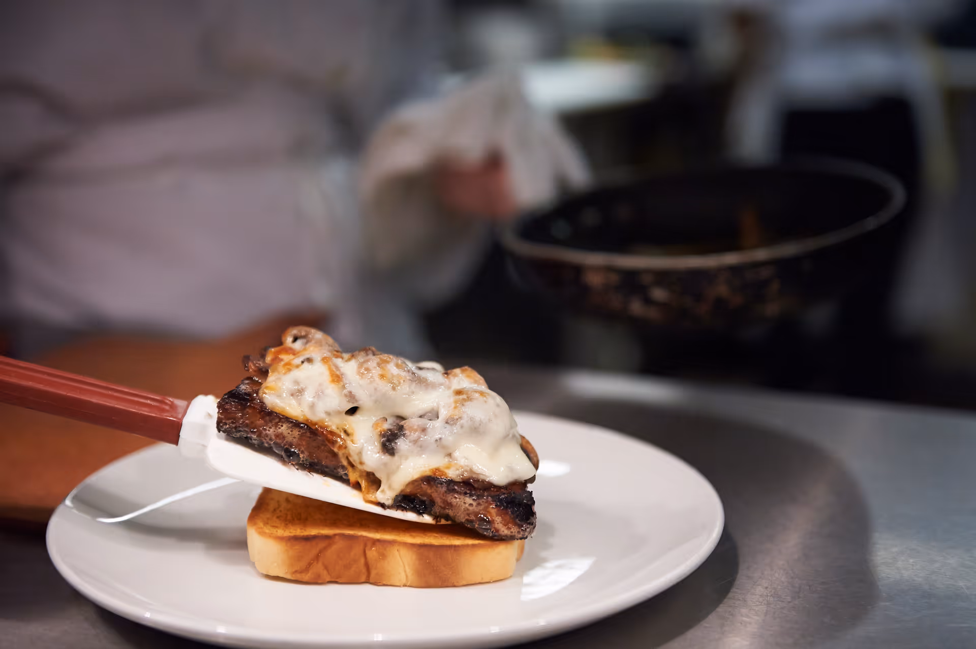 A spatula placing a cheesy grilled steak onto a slice of bread on a white plate in a kitchen.