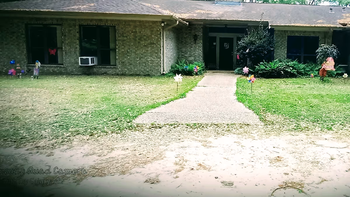 Front view of a single-story brick building with a paved walkway leading to the entrance. The lawn on either side of the walkway has colorful pinwheels and some bushes near the entrance. There are two windows visible on the left side, one with an air conditioning unit installed.