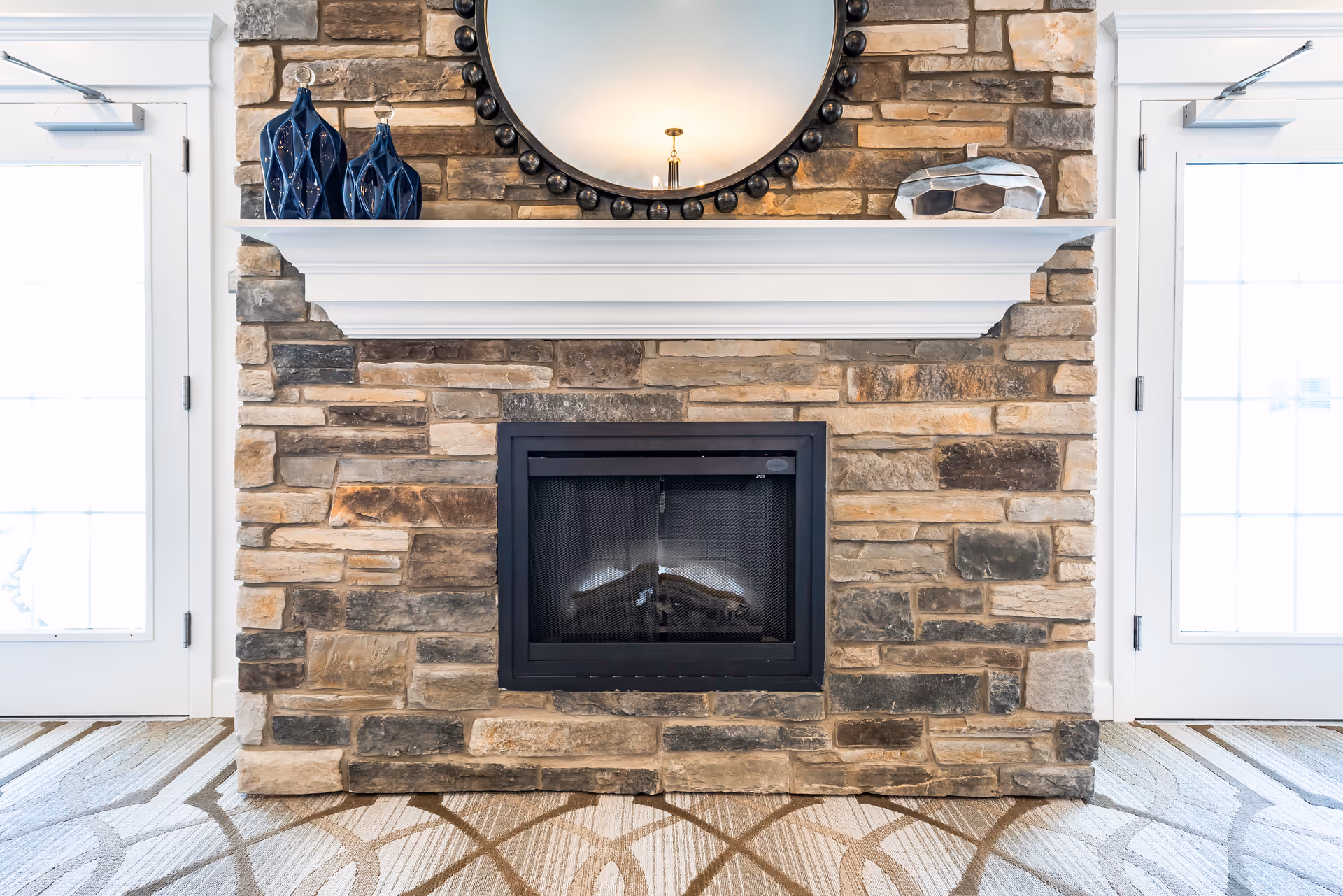 Stone fireplace with a white mantel, decorated with two blue vases and a geometric glass ornament. Above the mantel is a round mirror with a decorative frame. The fireplace is flanked by two white doors with glass panels, and the floor has a patterned carpet.