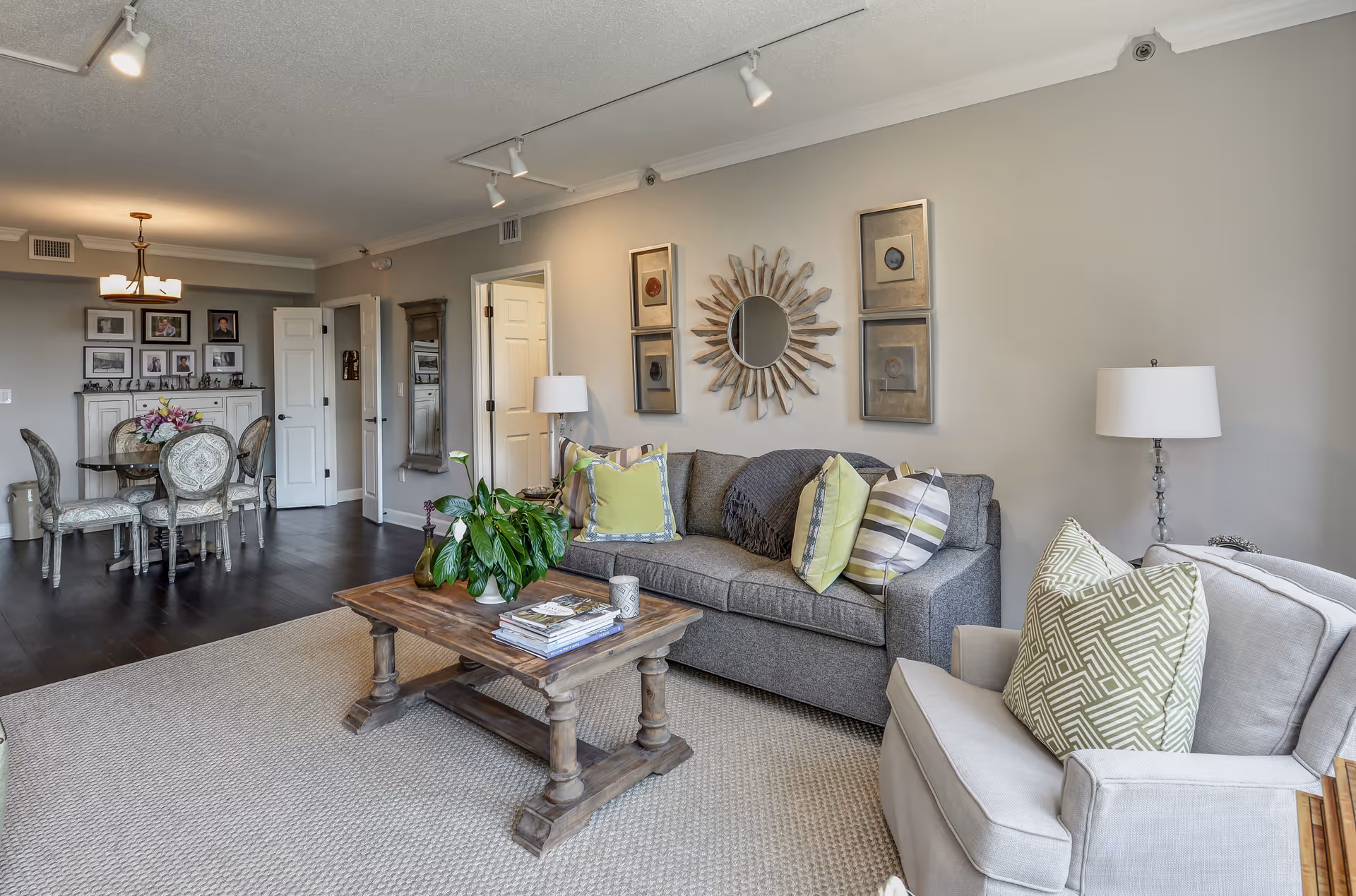 A cozy living room with a gray sofa adorned with patterned pillows, a wooden coffee table with books and a plant, and a beige armchair with a green patterned pillow. The wall behind the sofa features a decorative sunburst mirror and framed artwork. In the background, there is a dining area with a round table and four upholstered chairs, a chandelier, and a cabinet with framed photos and decorations.