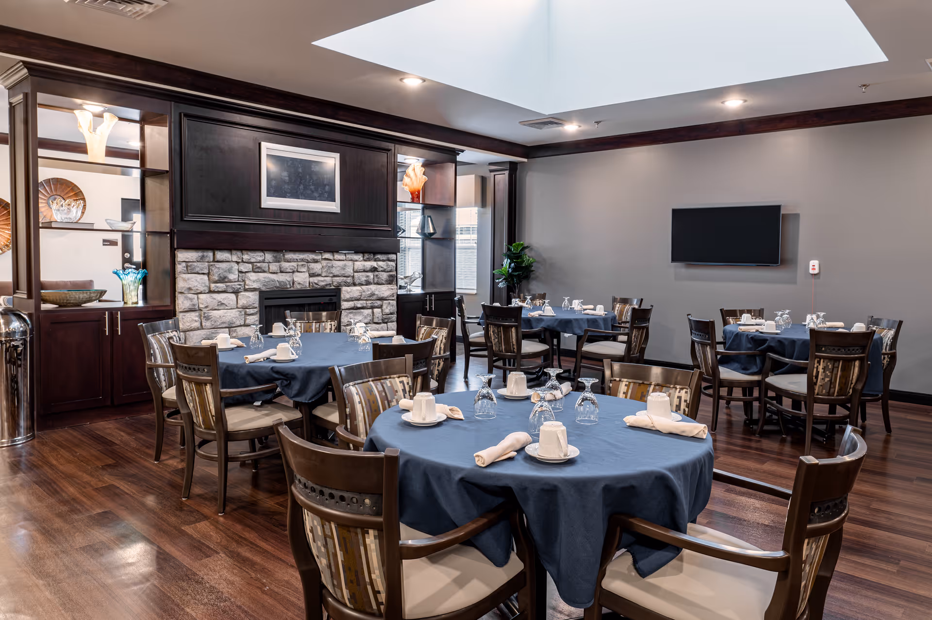 A dining room with multiple round tables covered with dark blue tablecloths, each set with white cups, napkins, and upside-down glasses. The room features wooden chairs with patterned cushions, a stone fireplace with dark wood cabinetry and shelves displaying decorative items, a flat-screen TV mounted on a gray wall, and a skylight in the ceiling providing natural light.