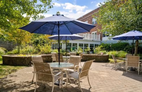 Outdoor patio area with round glass tables and beige chairs under large blue umbrellas, surrounded by greenery and trees, with a brick building in the background under a partly cloudy sky.