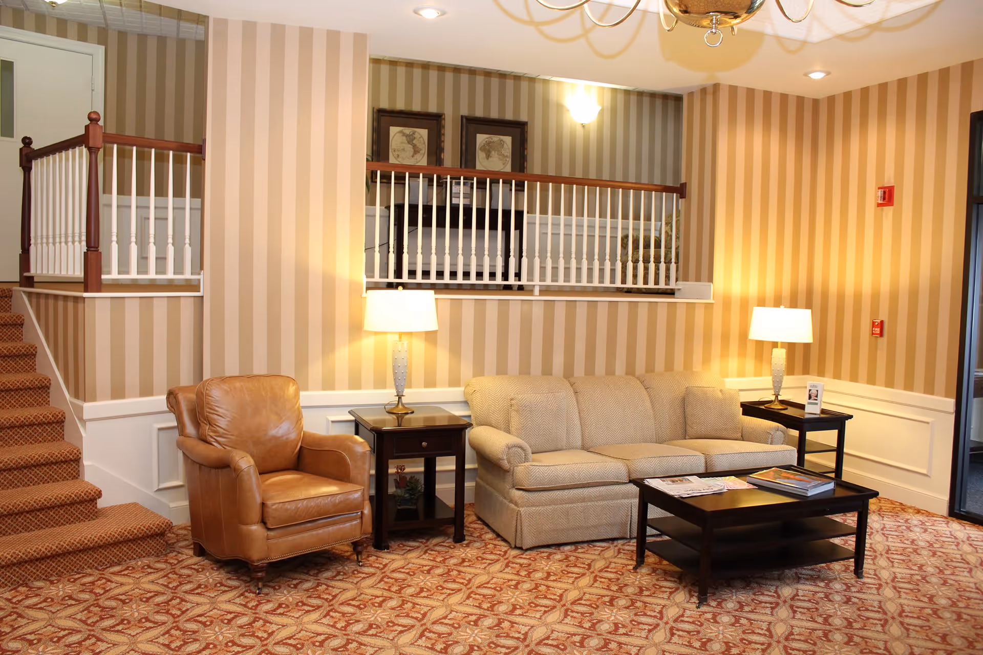 A cozy living room area with a beige sofa, a brown leather armchair, two side tables with lamps, a coffee table with magazines, and striped wallpaper. There is a staircase with carpet leading up to a railing and framed pictures on the wall above.