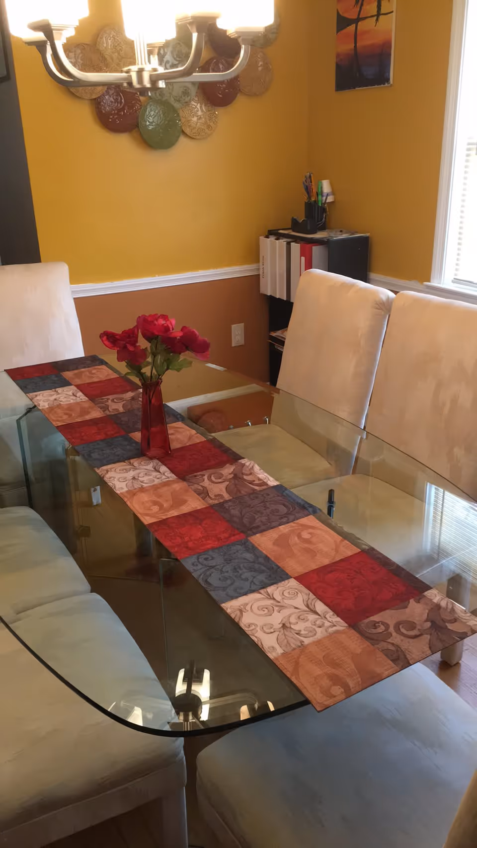 A dining room with a glass table covered by a colorful patchwork table runner and a red vase with red flowers in the center. The room has beige cushioned chairs around the table, yellow walls with decorative plates hanging, and a window letting in natural light.