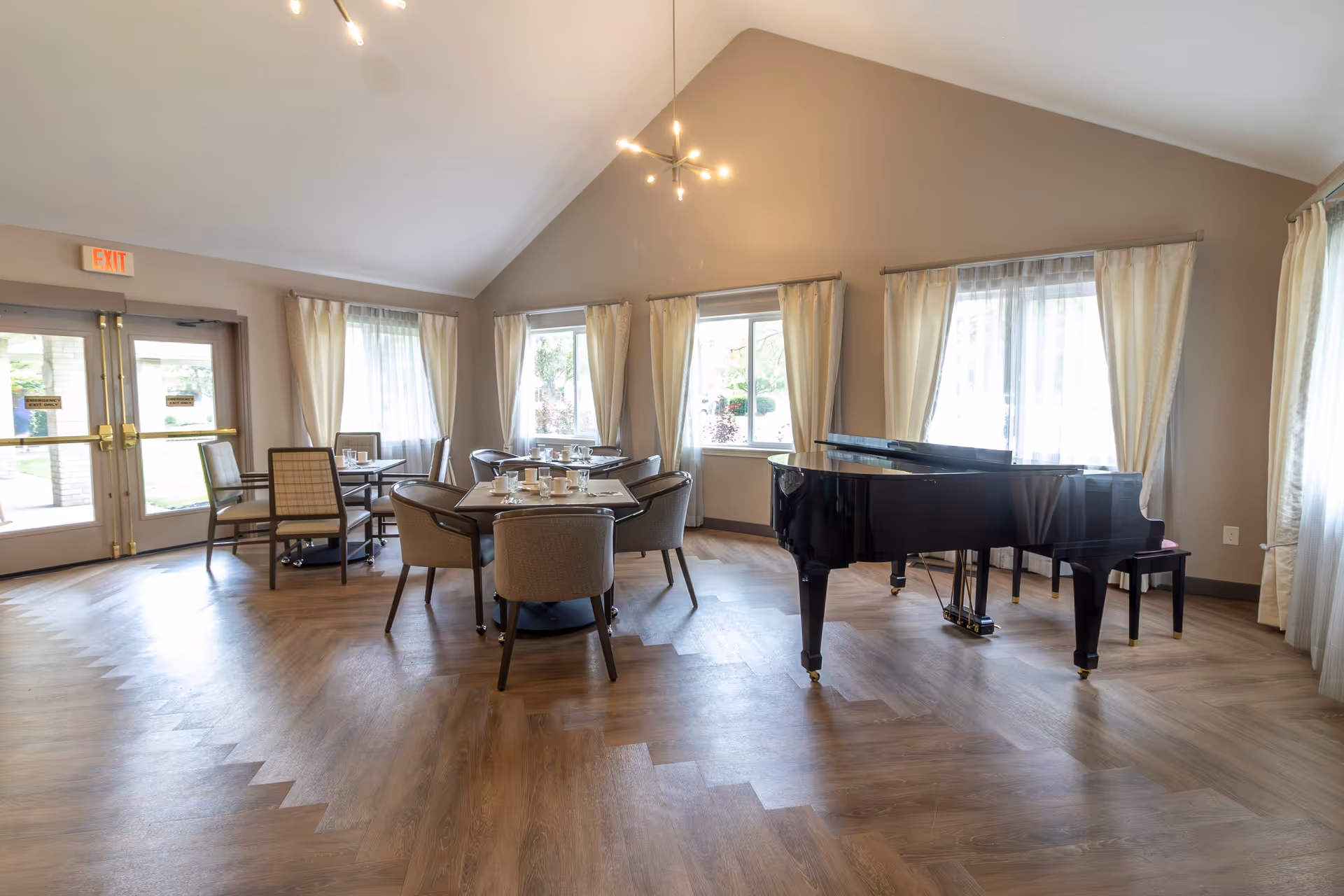 A bright room with large windows covered by light curtains, featuring a black grand piano on the right side and several dining tables with chairs arranged around them. The room has a high ceiling with modern light fixtures and wood-patterned flooring. Double glass doors with exit signs are visible on the left side.
