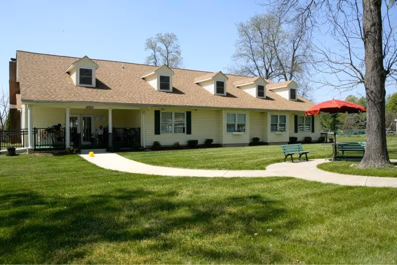 Single-story building with a beige exterior and brown roof, featuring four dormer windows. A concrete pathway leads to the entrance porch where two people are seated. The surrounding area has well-maintained green grass, a large tree, two green benches, and a table with a red umbrella.