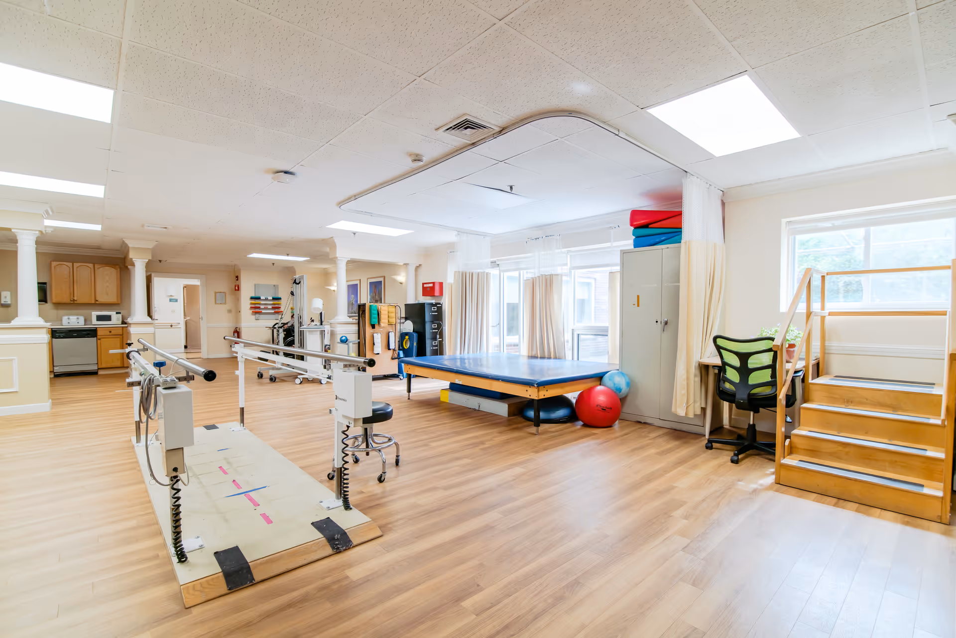 A spacious rehabilitation or physical therapy room with wooden flooring, parallel bars for walking exercises, a blue therapy mat, exercise balls, a small desk with a green office chair, and wooden stairs for mobility training. The room is well-lit with large windows and overhead fluorescent lights.