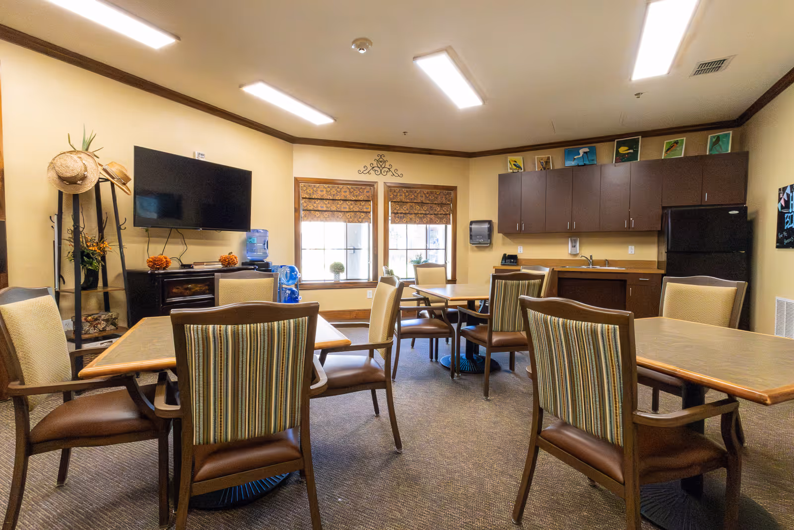 Communal dining/activity room with tables and chairs, a wall-mounted TV, kitchenette cabinets, and windows.