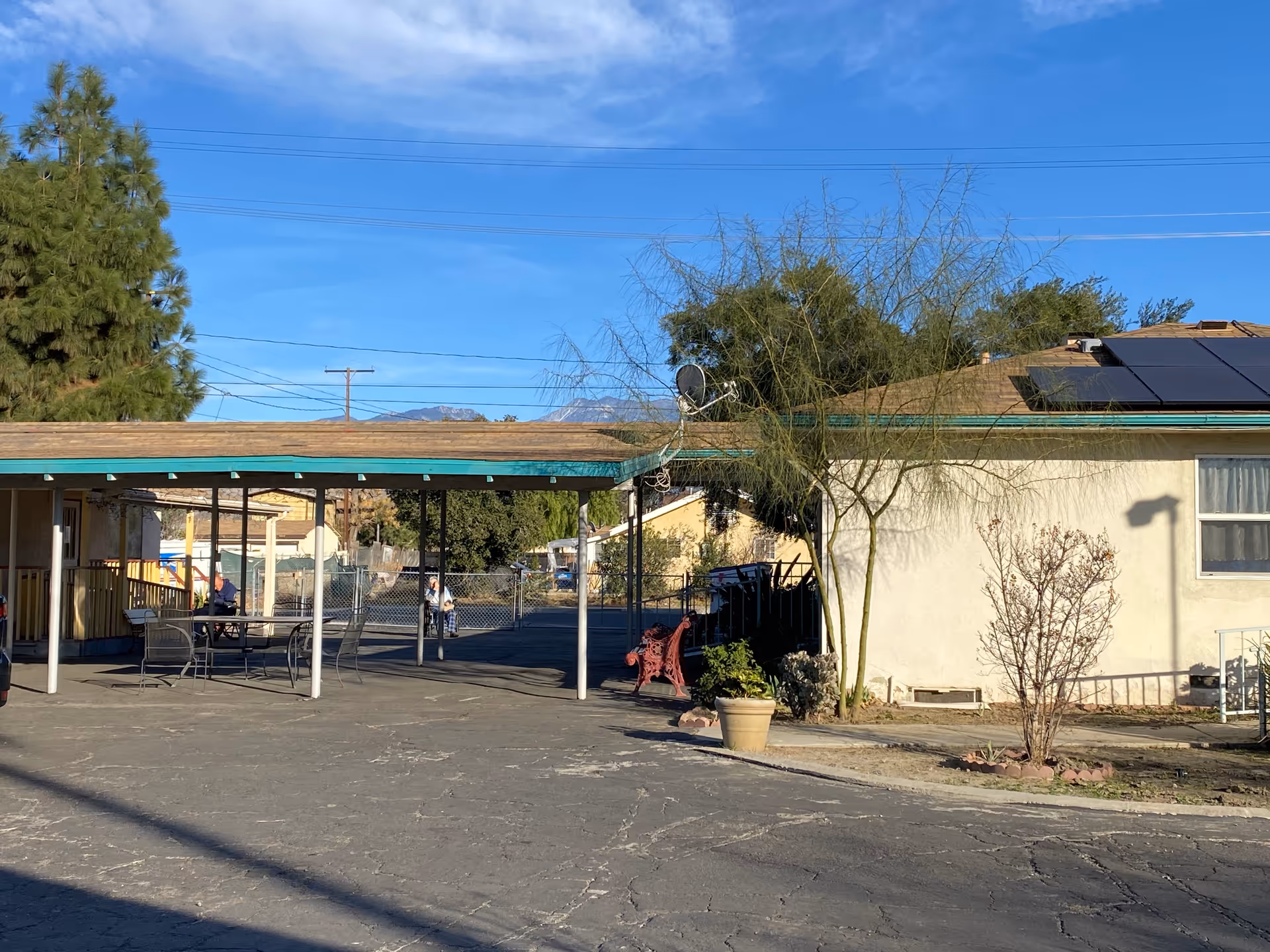 Outdoor area of Azzur Assisted Living facility showing a covered patio with tables and chairs, a few people sitting and walking, trees, and a building with solar panels on the roof under a clear blue sky.