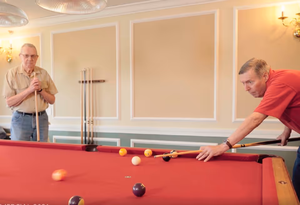 Two elderly men playing pool in a well-lit room with beige walls and decorative molding. One man is preparing to take a shot while the other watches, holding a cue stick. Several pool cues are mounted on the wall behind them.