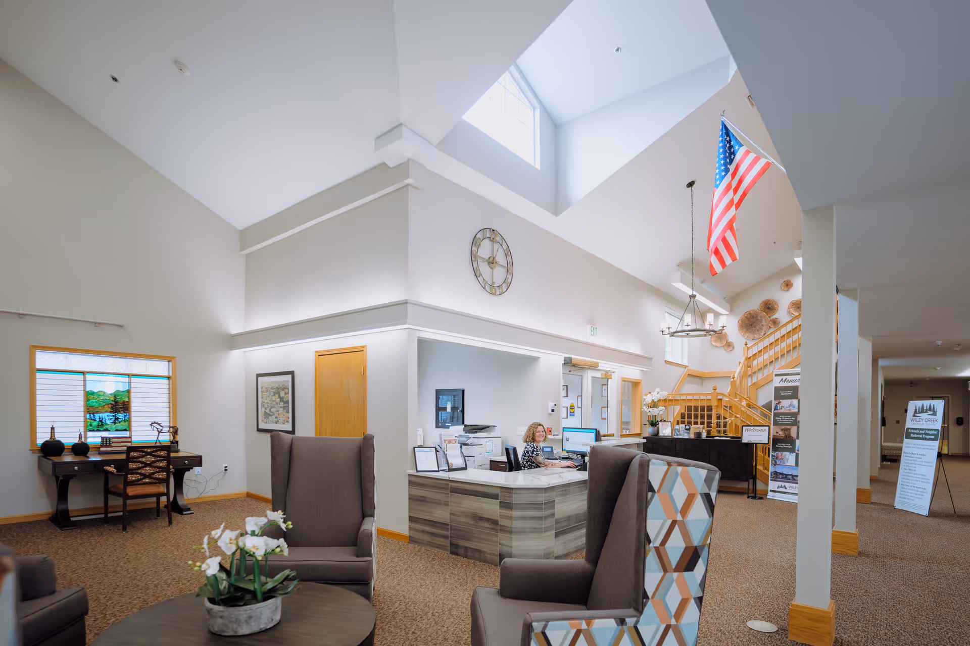 Interior view of a senior living facility reception area with high ceilings and skylights. There is a reception desk with a staff member sitting behind it, two armchairs with patterned upholstery, a round table with a flower arrangement, a desk with a stained glass window, and an American flag hanging from the ceiling. A staircase and informational signs are visible in the background.