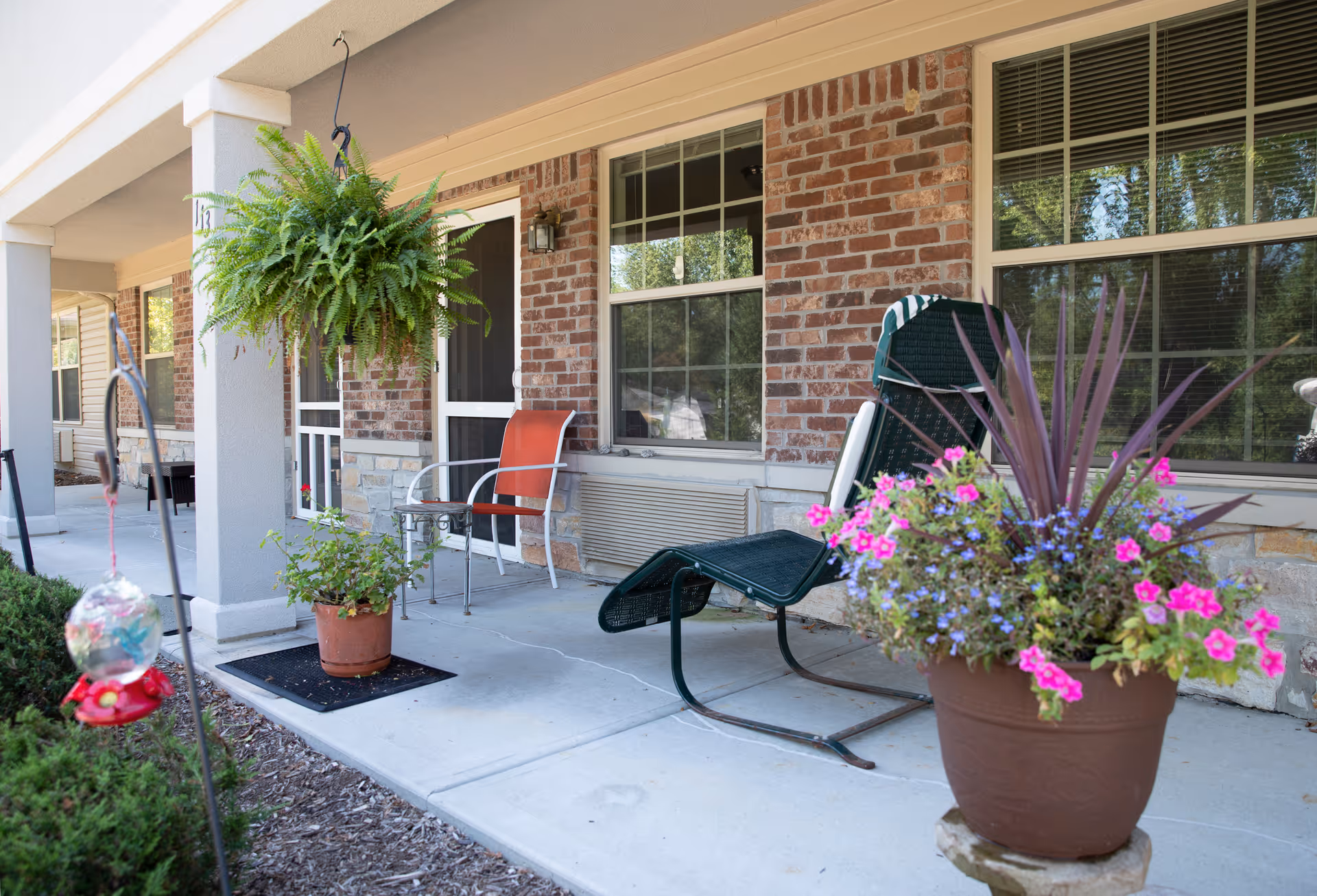 Front porch with potted flowers, a hanging fern, and chairs outside a brick-faced assisted living building.