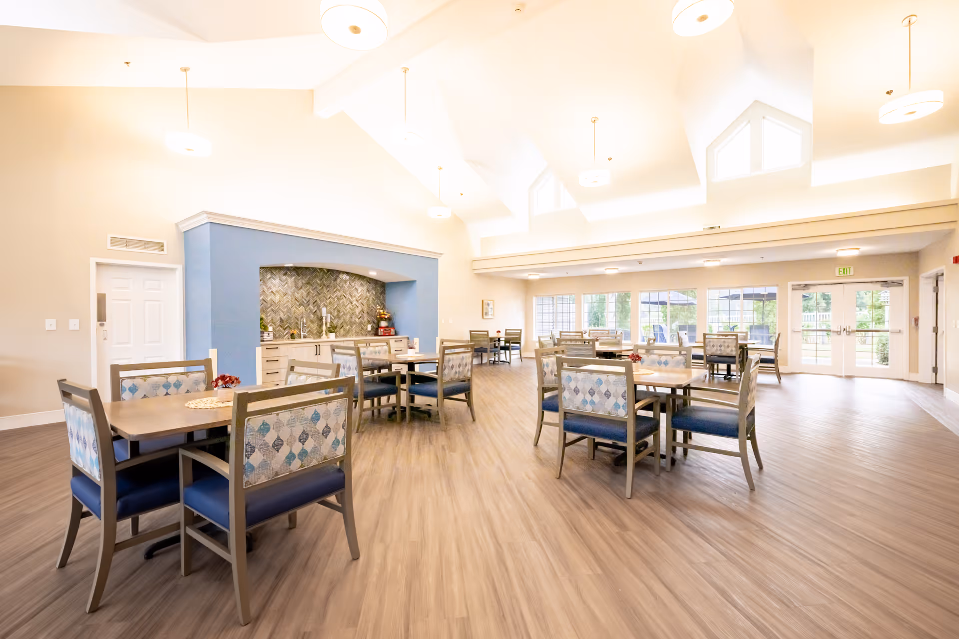 A spacious, well-lit dining room with multiple tables and chairs arranged neatly. The room features high ceilings with modern hanging light fixtures, large windows allowing natural light to fill the space, and a blue accent wall with a built-in counter and decorative backsplash. The floor is covered with light wood-style flooring.