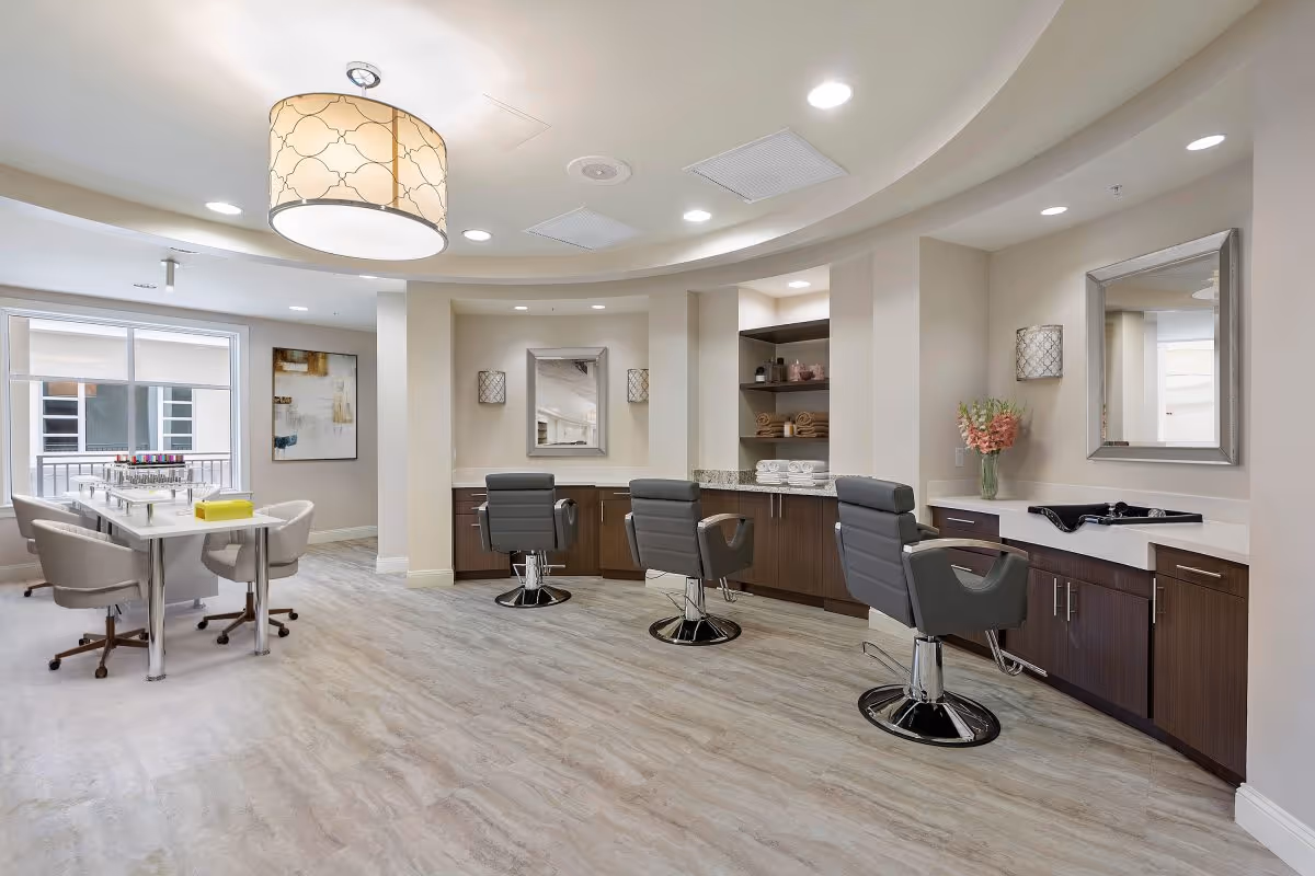 Bright modern salon-style common room with styling chairs, mirrors, and a manicure table.