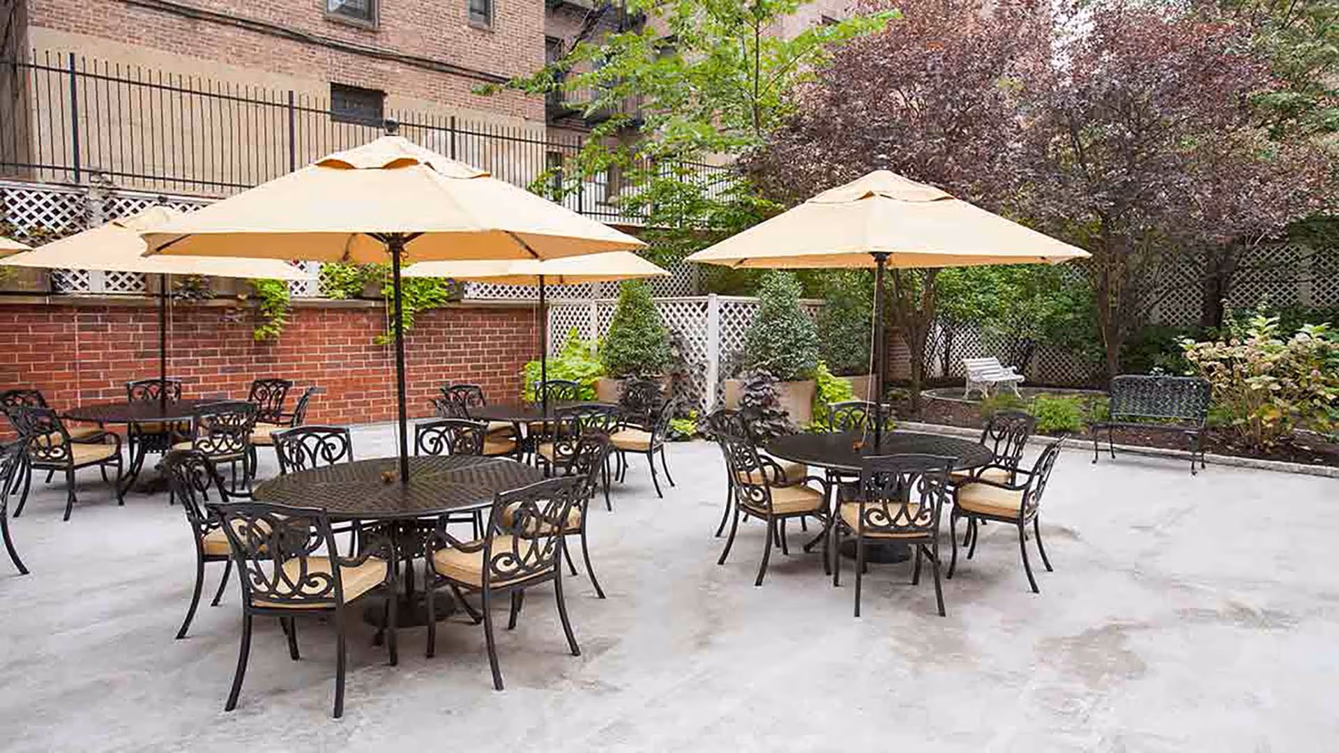 Outdoor patio area with several round black metal tables and matching chairs with beige cushions. Each table has a large beige umbrella providing shade. The patio is surrounded by brick walls, lattice fencing, and various green plants and trees.