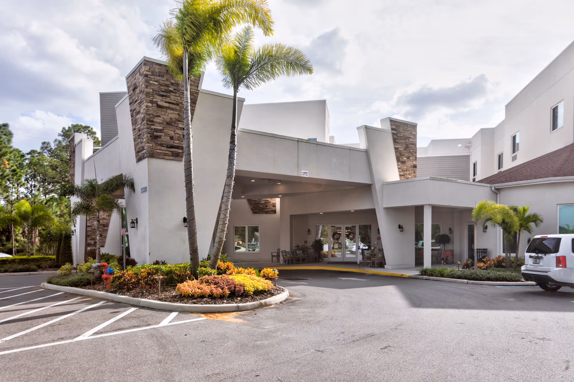 Exterior view of the entrance to a senior living facility named Discovery Village At Westchase, featuring a covered drop-off area, palm trees, landscaped plants, and a parking area with a white SUV parked nearby.