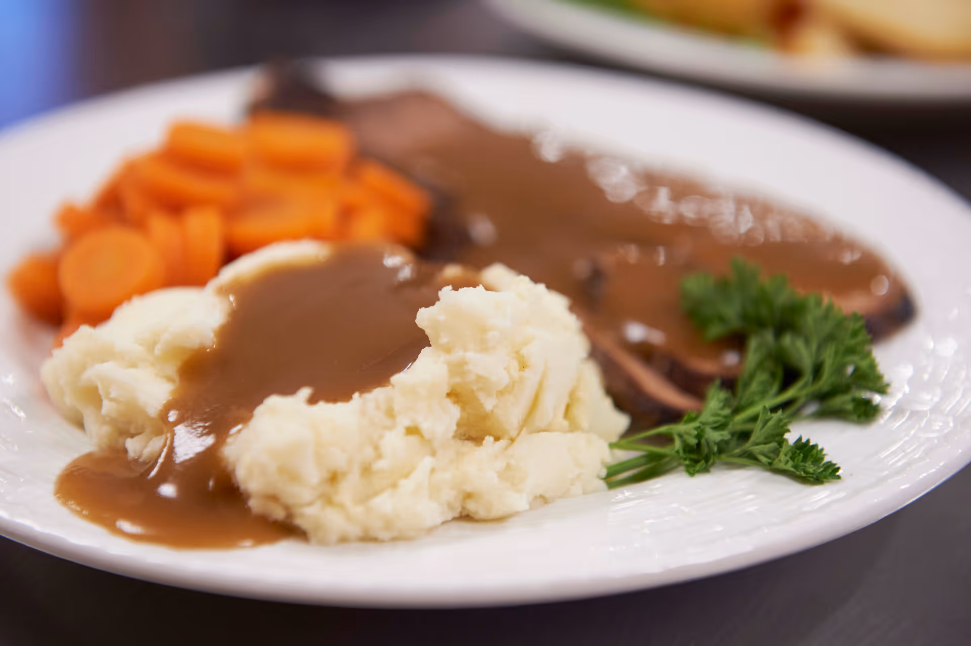 A white plate with mashed potatoes topped with brown gravy, sliced roast beef with brown gravy, cooked sliced carrots, and a sprig of parsley as garnish.