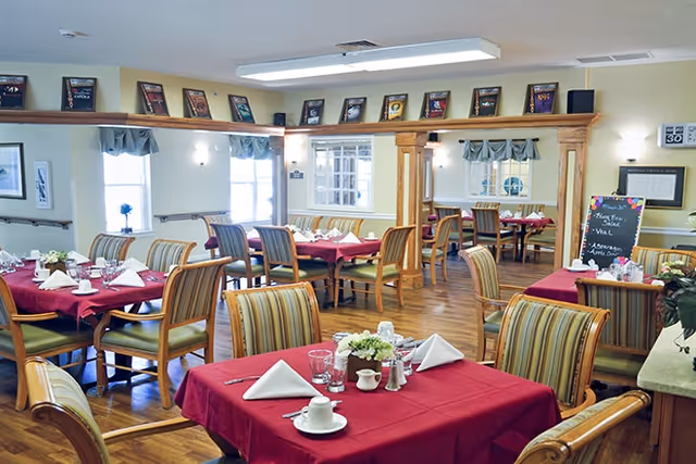 A dining room with multiple tables covered in red tablecloths, each set with white napkins, cups, glasses, and small flower arrangements. The room has wooden floors, striped cushioned chairs, large windows with valances, and framed pictures displayed on a ledge near the ceiling.