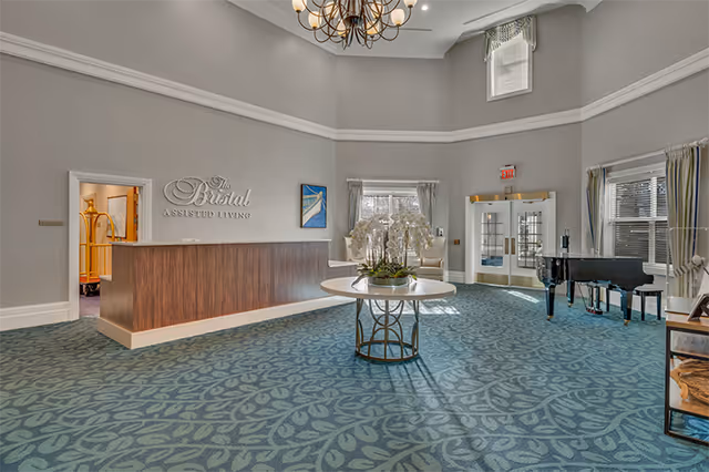 Spacious assisted living lobby with a reception desk, central round table with flowers, and a grand piano.