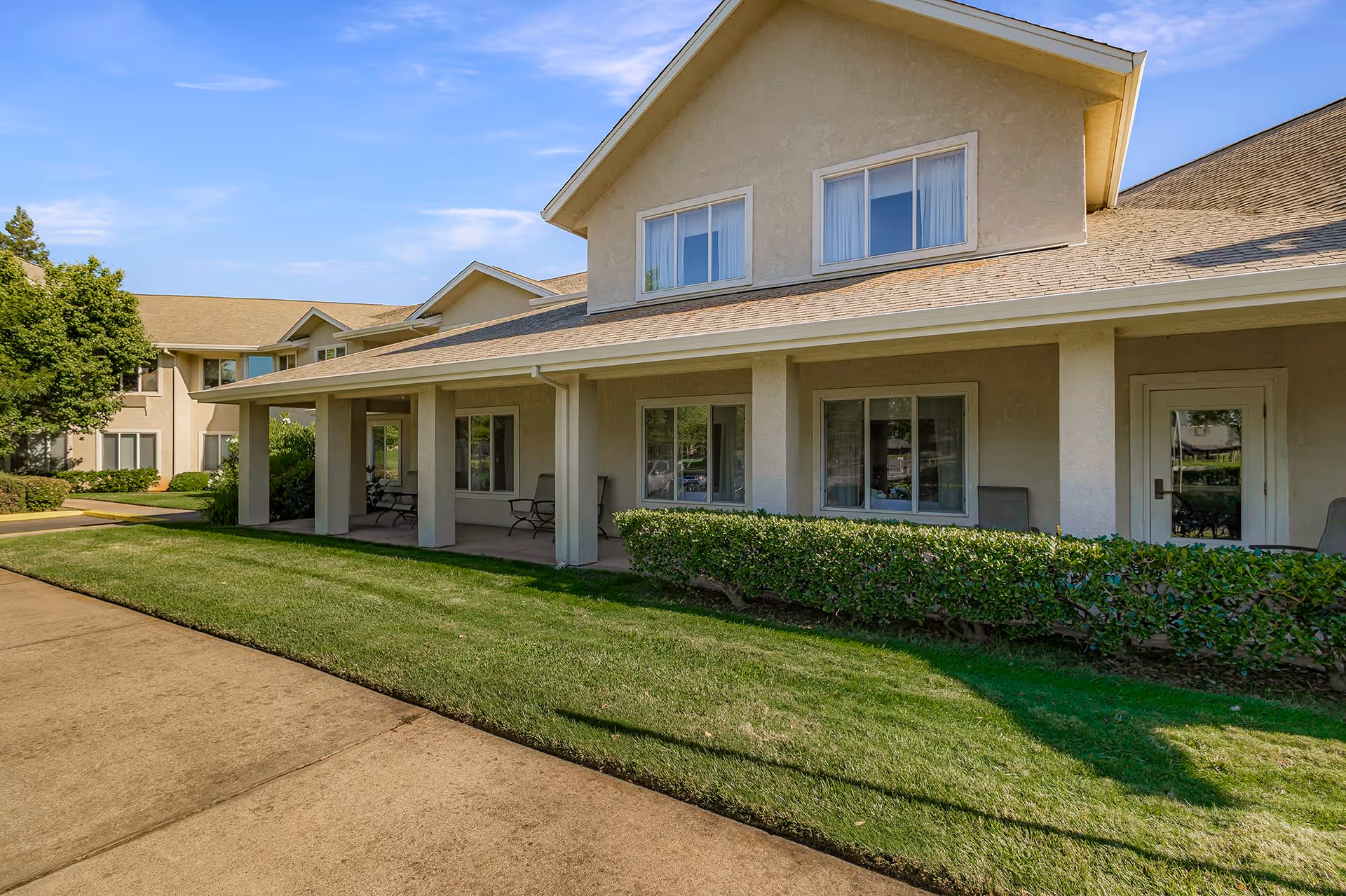 Exterior view of a beige two-story building with a covered porch, several windows, and a door. There are green bushes and a well-maintained lawn in front, with a concrete walkway alongside the building under a clear blue sky.
