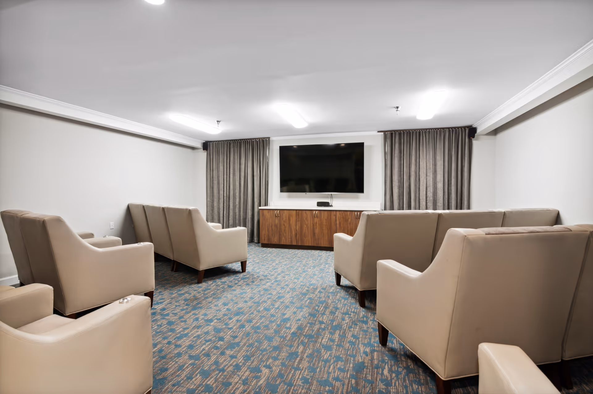 A small media room with beige armchairs arranged in two rows facing a large flat-screen TV mounted on the wall above a wooden cabinet. The room has gray curtains on either side of the TV and a patterned blue carpet.