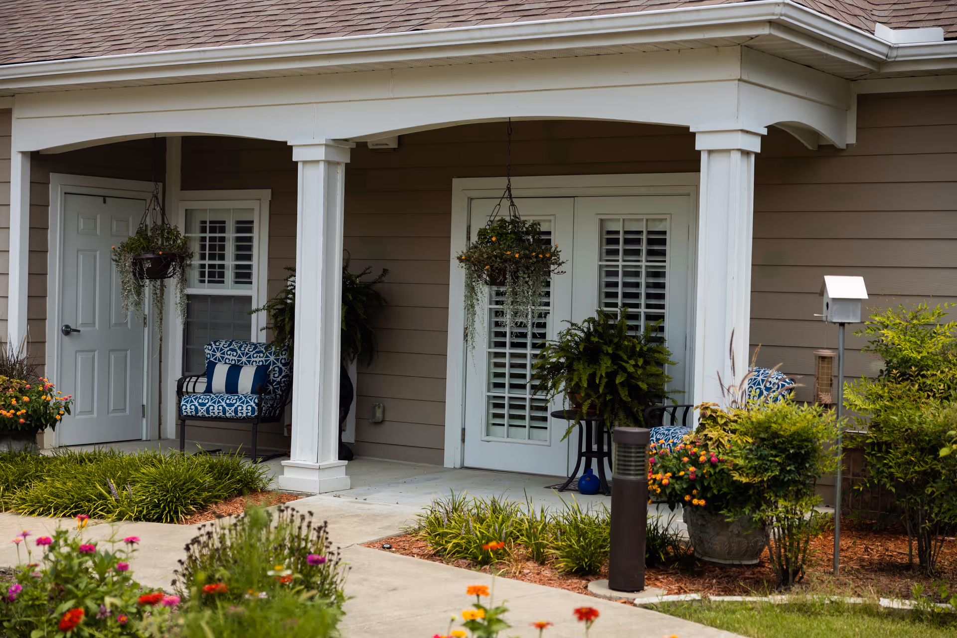 Covered front porch with white columns, double French doors, patterned outdoor chairs, hanging plants and a landscaped walkway.