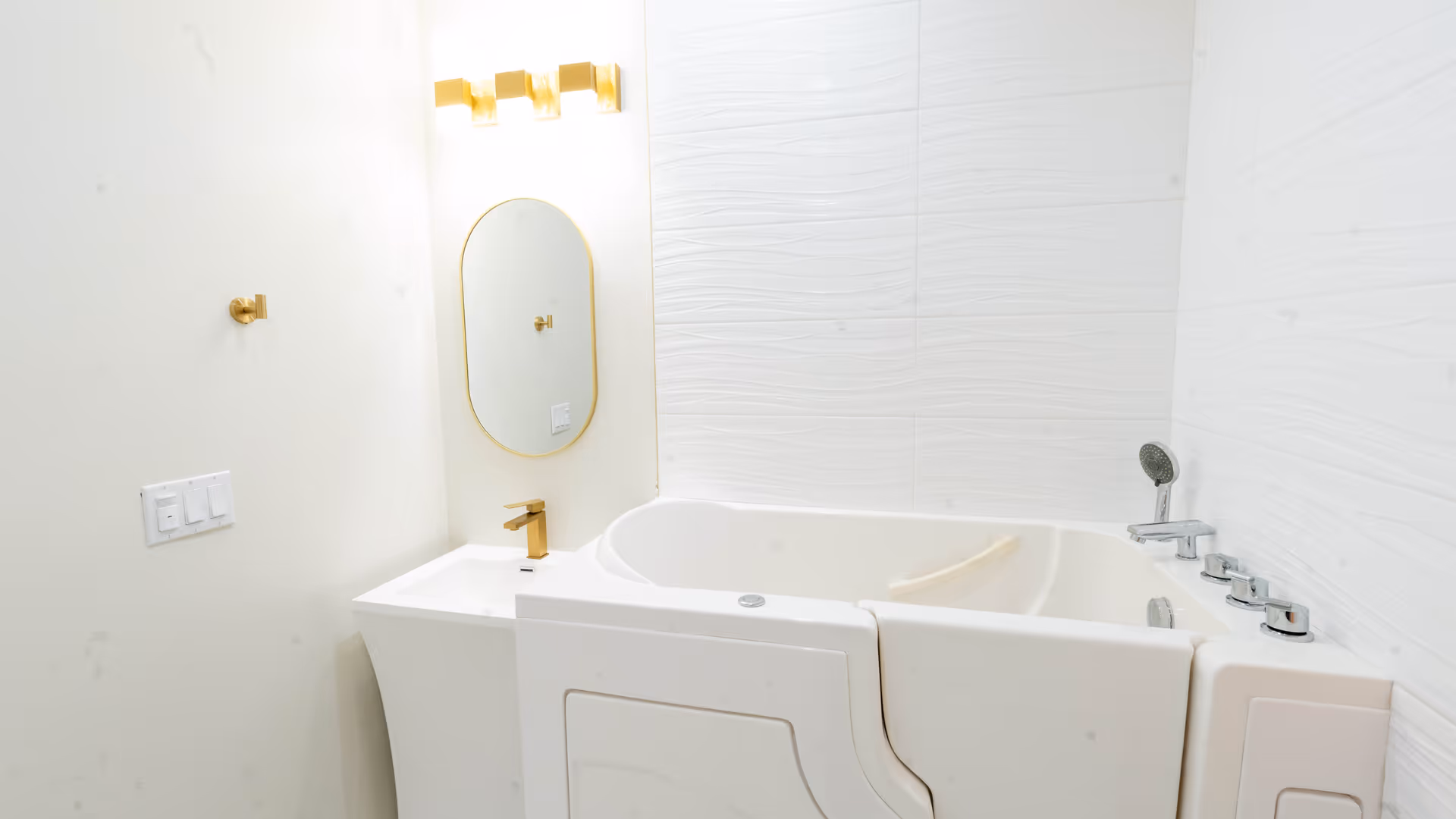 A modern bathroom featuring a walk-in bathtub with multiple faucet controls and a handheld showerhead. The walls have white textured tiles, and there is a small white sink with a gold faucet and an oval mirror with a gold frame above it. A gold light fixture is mounted above the mirror.