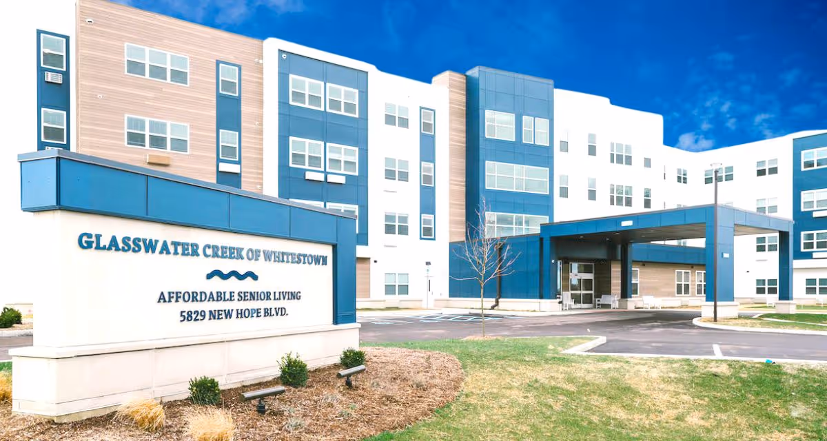 Exterior view of Glasswater Creek of Whitestown, a multi-story senior living facility with a modern design featuring blue and beige panels. A large sign in front reads 'Glasswater Creek of Whitestown, Affordable Senior Living, 5829 New Hope Blvd.' The building has multiple windows and a covered entrance with a driveway and parking spaces.